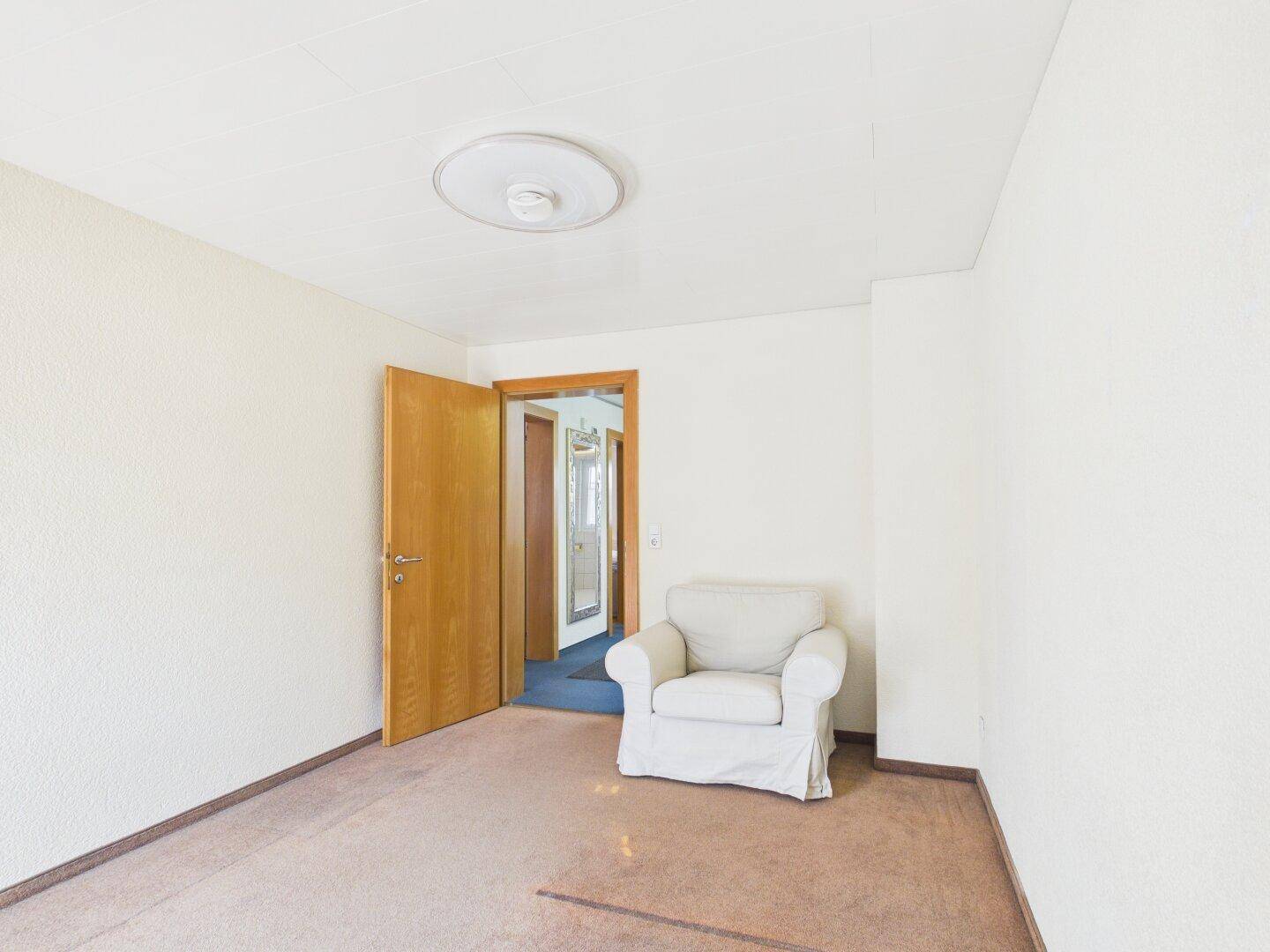 Helles Zimmer mit Teppichboden und Sessel, bereit zur individuellen Gestaltung.