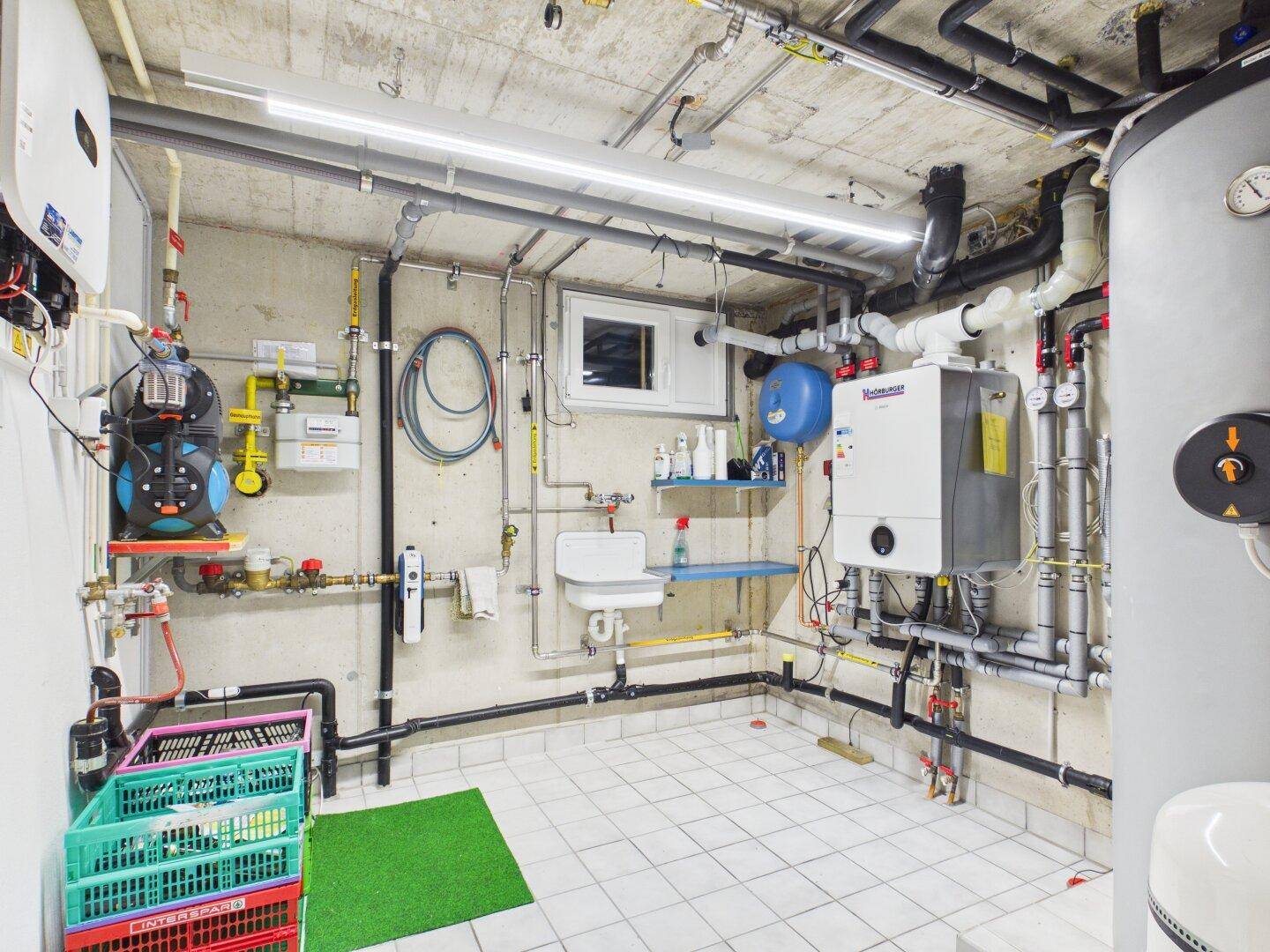 Technikraum mit Heizungsanlage, Warmwasserspeicher und zahlreichen Leitungen.