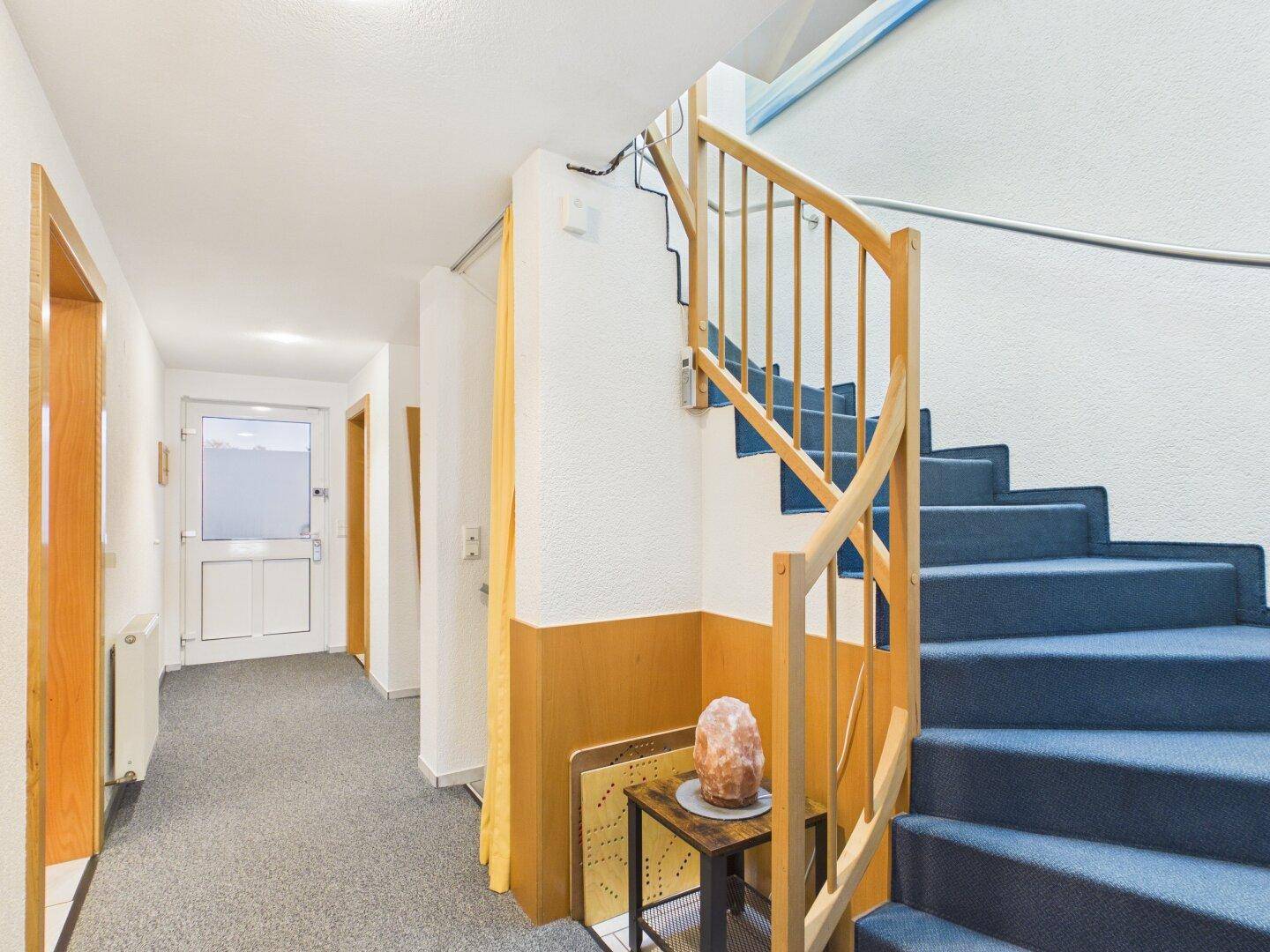 Flur mit blauer Teppichtreppe, Holzgeländer und Zugang zum Eingangsbereich.