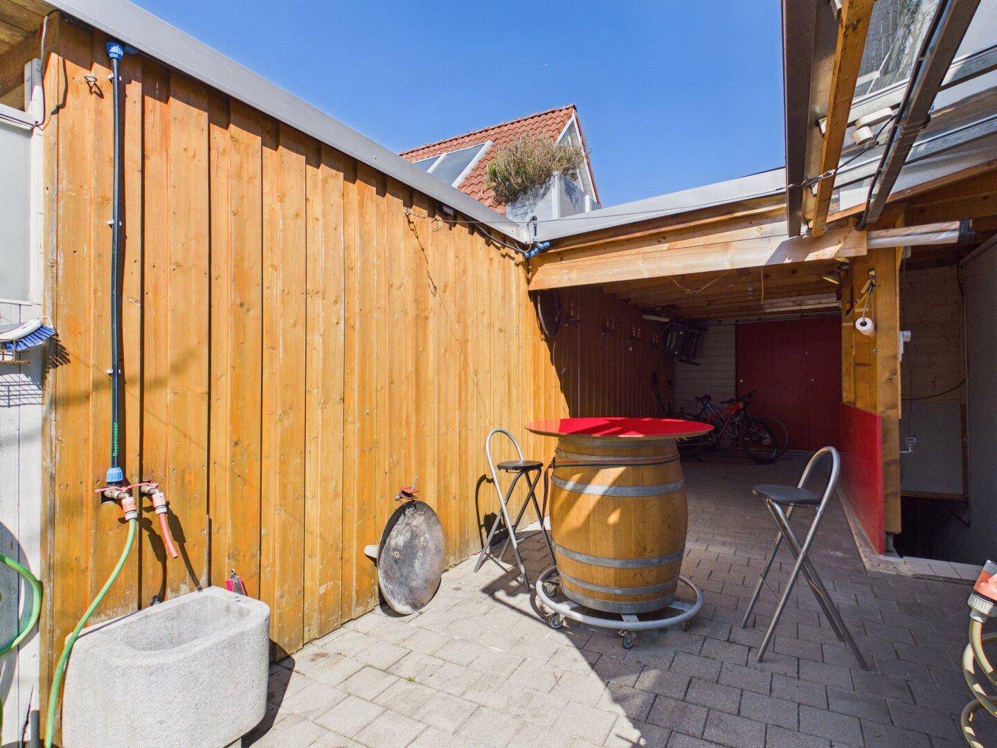 Gepflasterter Außenbereich mit Holzwand, Stehtisch und Zugang zu einem überdachten Gang.