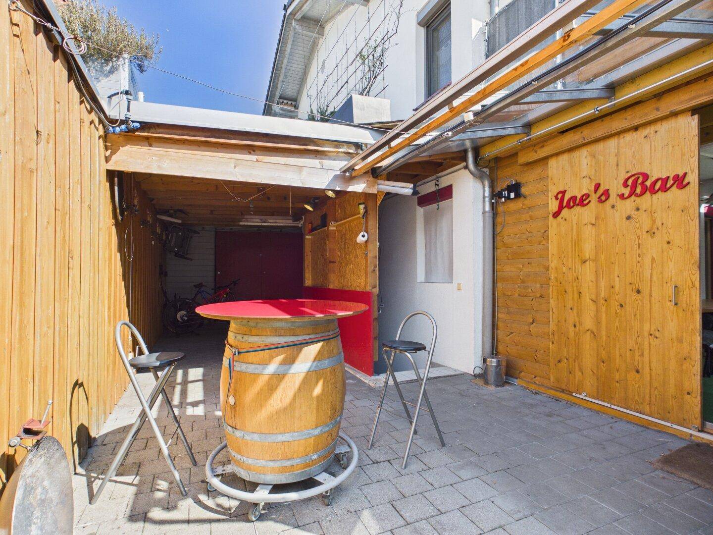 Außenbereich mit Holzwand, Stehtisch und "Joe's Bar" Schriftzug.