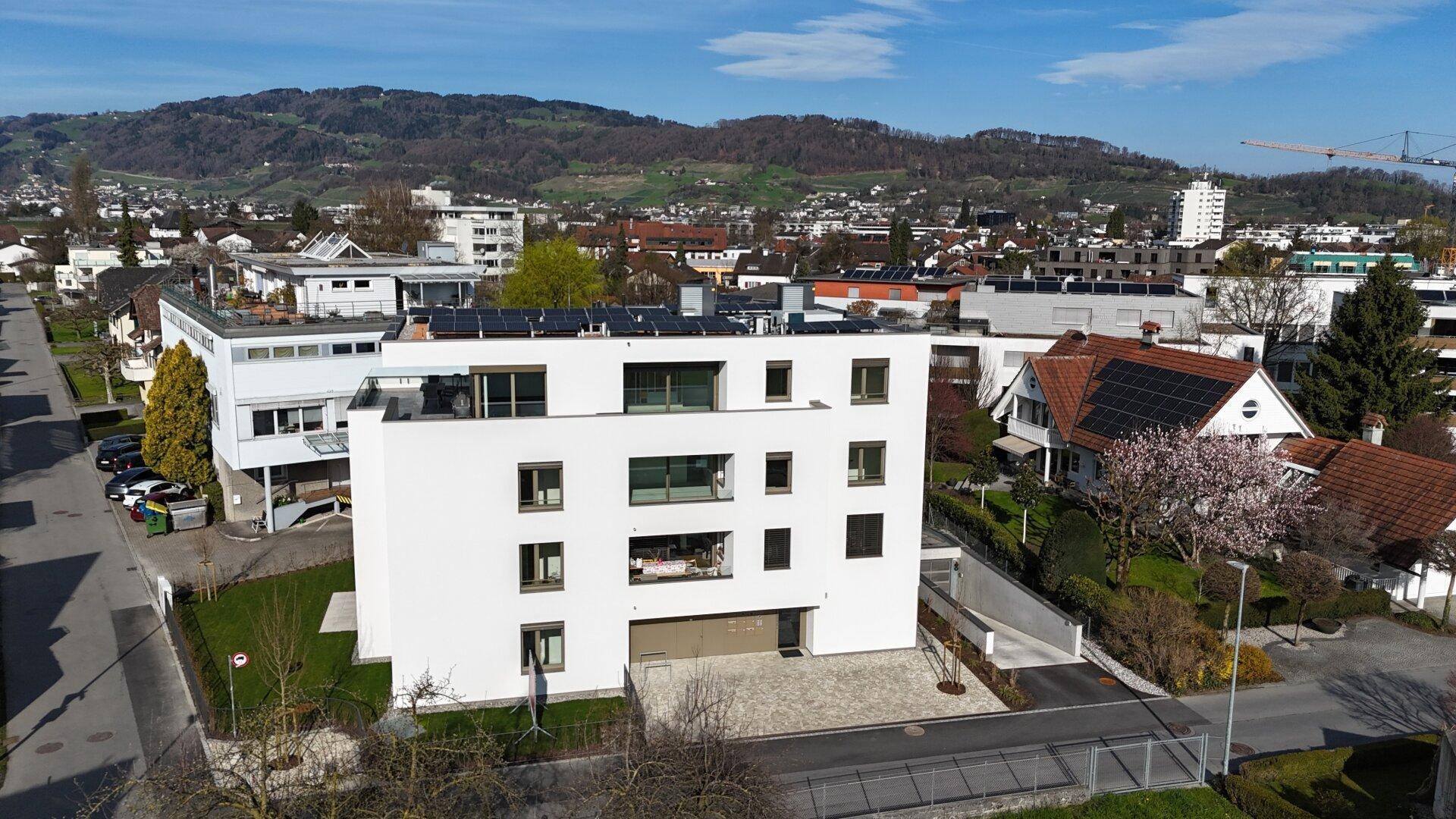 Modernes Wohngebäude mit weißer Fassade und Photovoltaikanlage auf dem Dach, umgeben von grüner Landschaft.