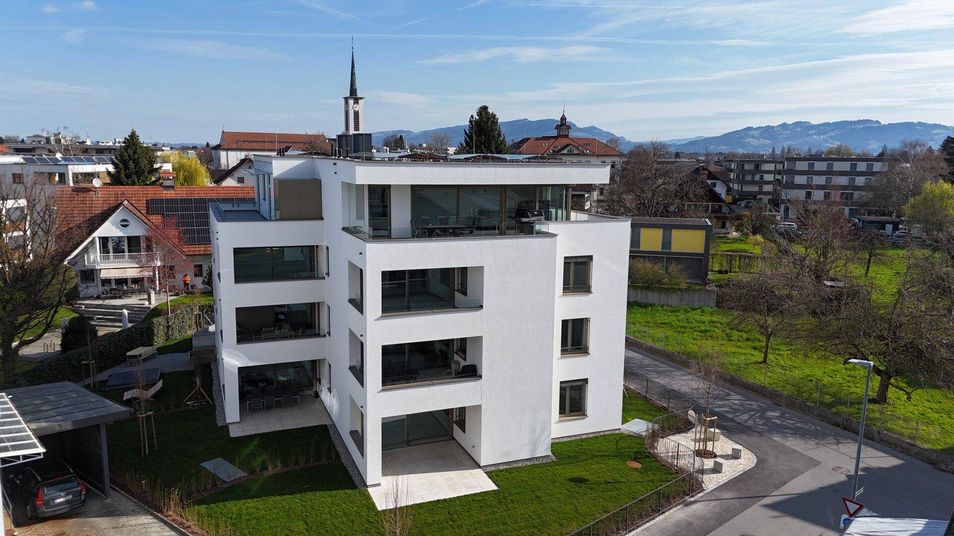 Modernes Gebäude mit weißer Fassade, großen Glasfronten und mehreren Balkonen sowie einer Dachterrasse.