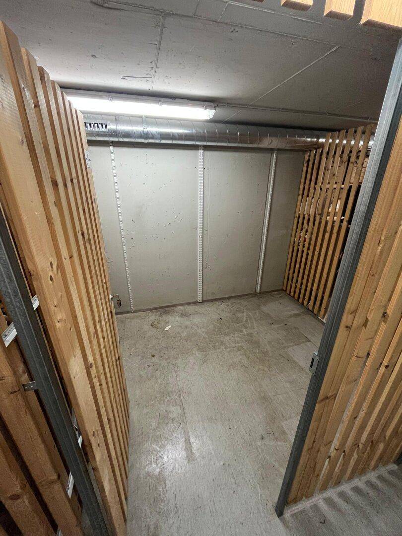 Leeres Kellerabteil mit Betonboden und Holzlattenwänden, ausgestattet mit einfacher Beleuchtung und sichtbaren Rohren.