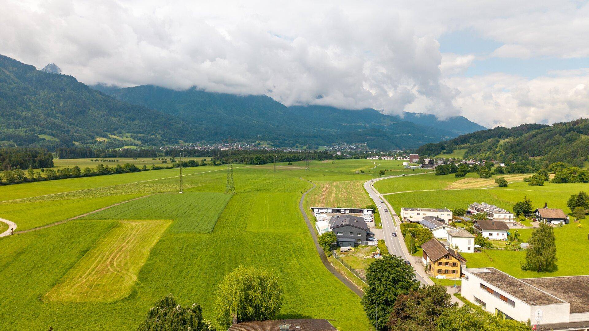 Panoramablick über ein grünes Tal mit Häusern, Feldern und einer Straße, umgeben von Bergen.