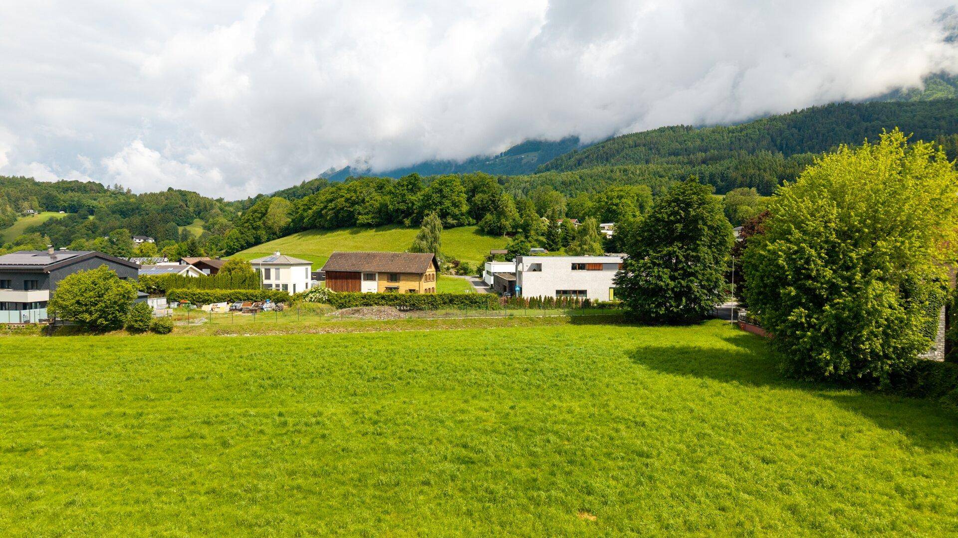 Weitläufige grüne Wiesen mit vereinzelten Häusern und bewaldeten Bergen im Hintergrund.
