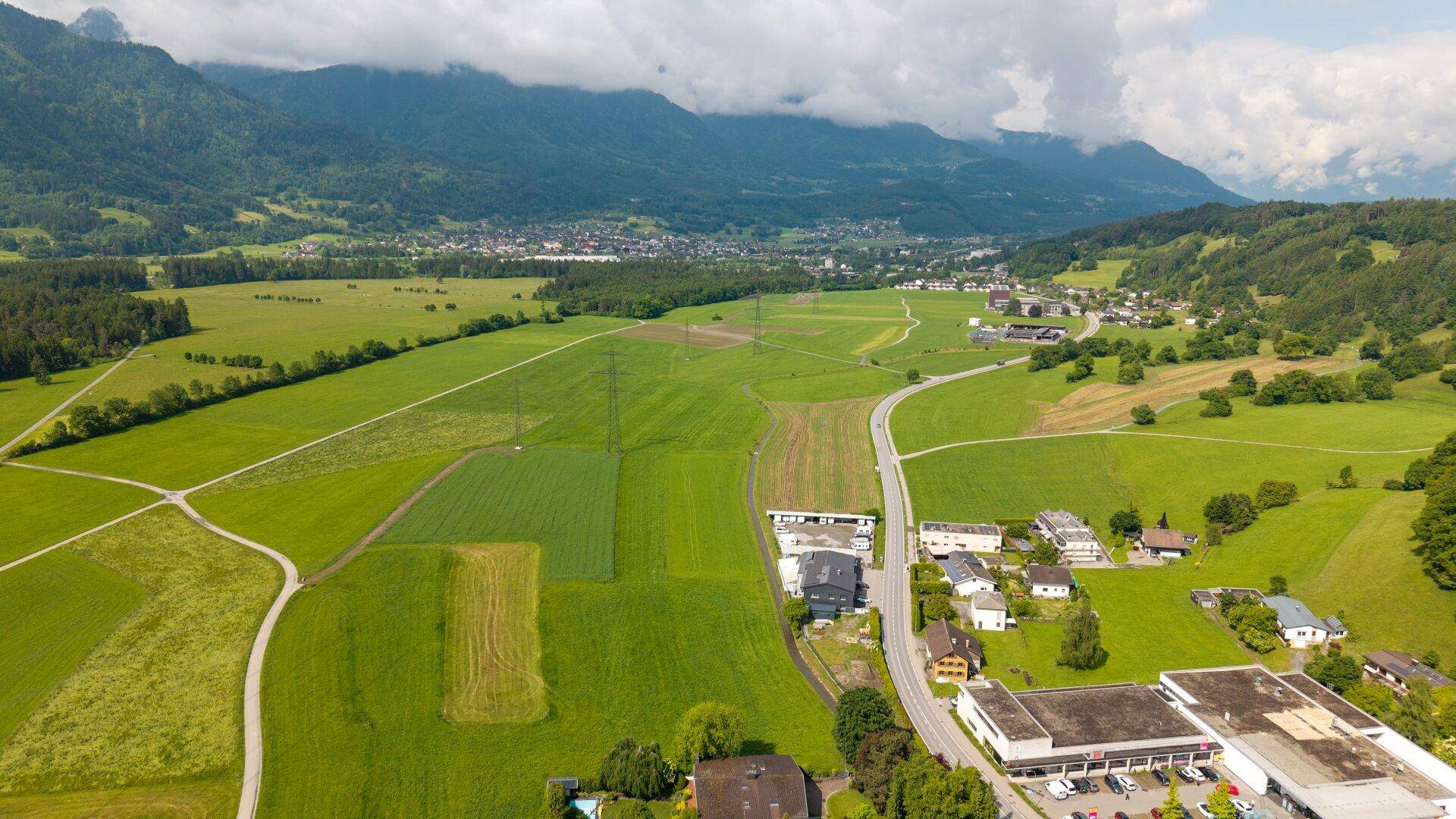 Breiter Blick über ein grünes Tal mit Feldern, Häusern und einer Straße, umrahmt von Bergen.