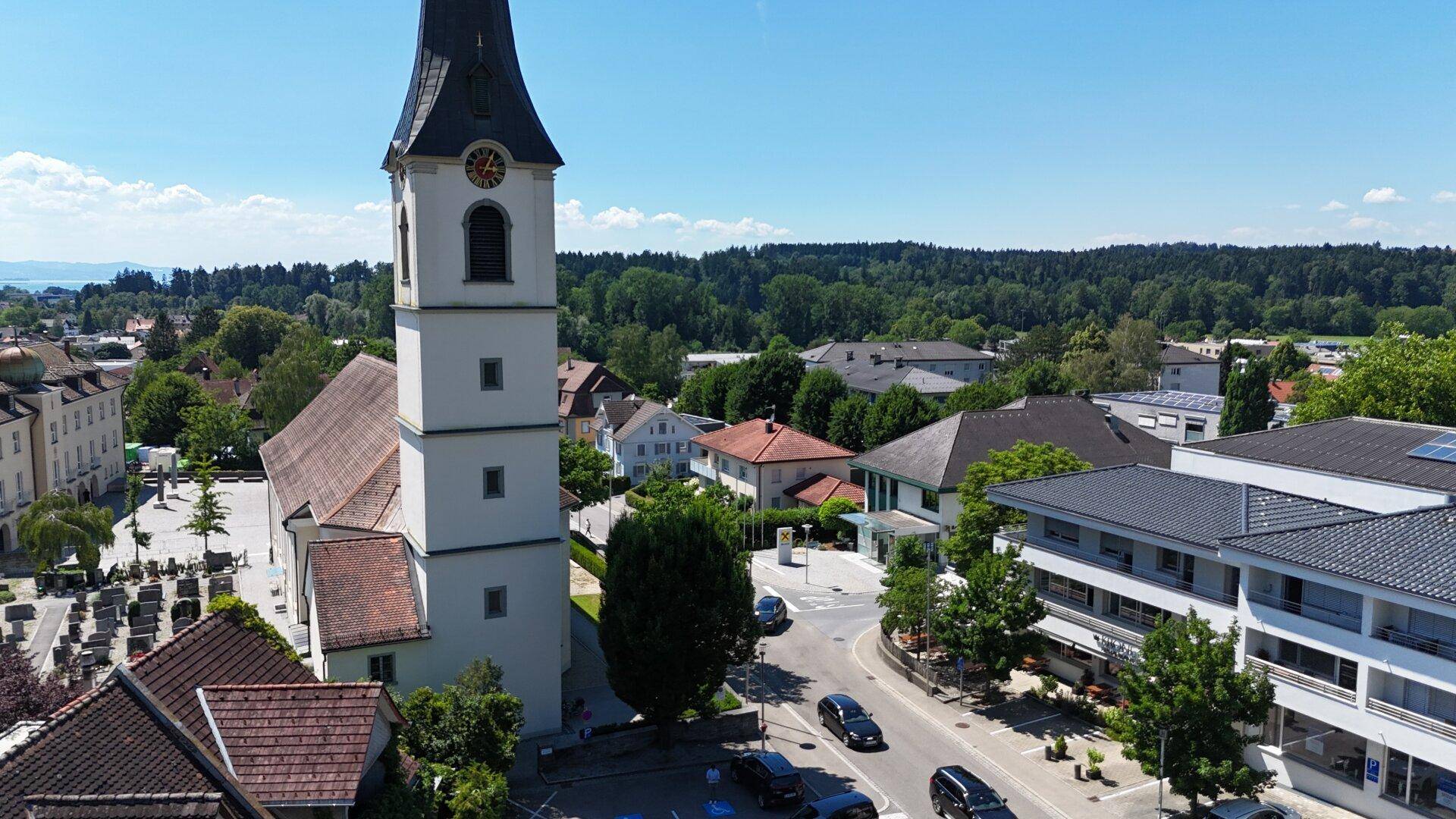 Luftaufnahme einer Stadt mit einer Kirche, umliegenden Gebäuden und einem See im Hintergrund unter blauem Himmel.