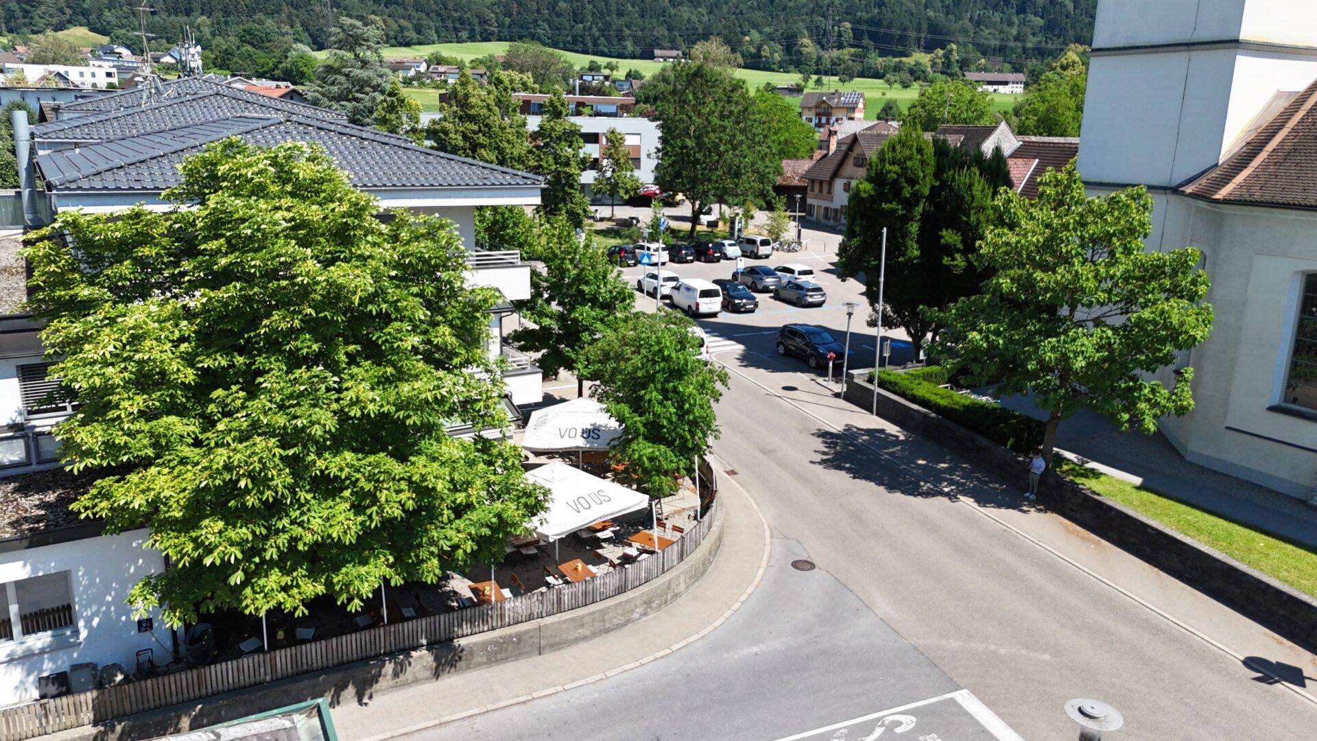 Vogelperspektive auf eine belebte Straßenecke mit einem Restaurant, Außenterrasse und umliegenden Gebäuden vor einer Bergkulisse.