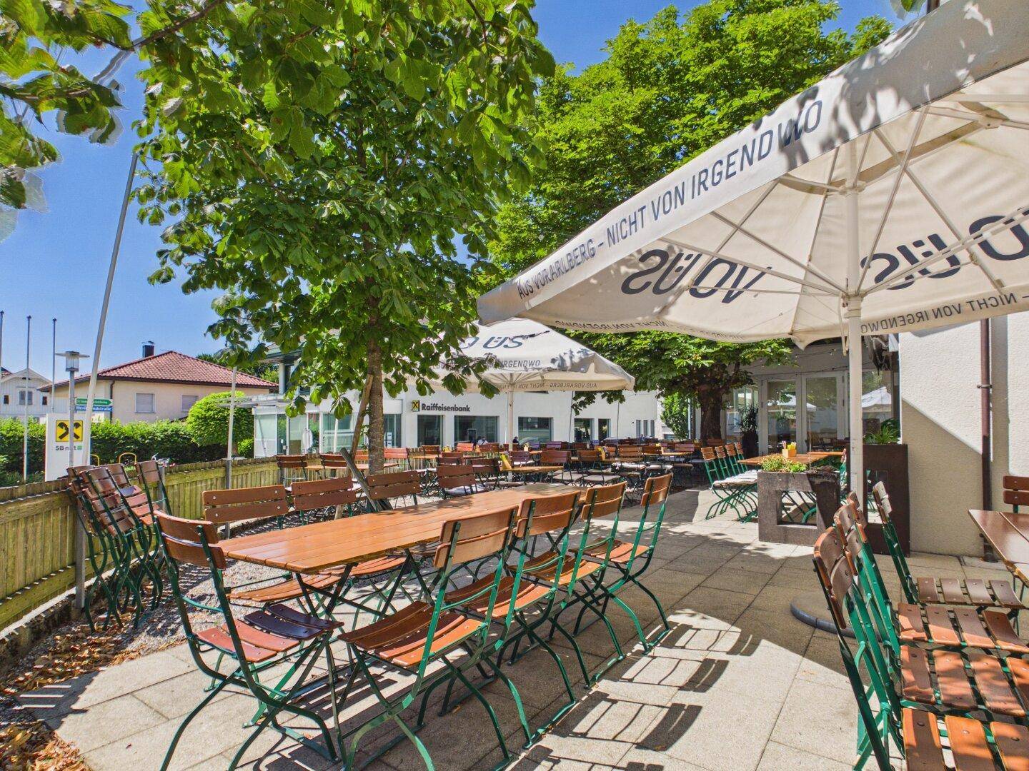 Detailansicht einer einladenden Restaurantterrasse mit Holztischen und grünen Stühlen, beschattet von großen Bäumen.