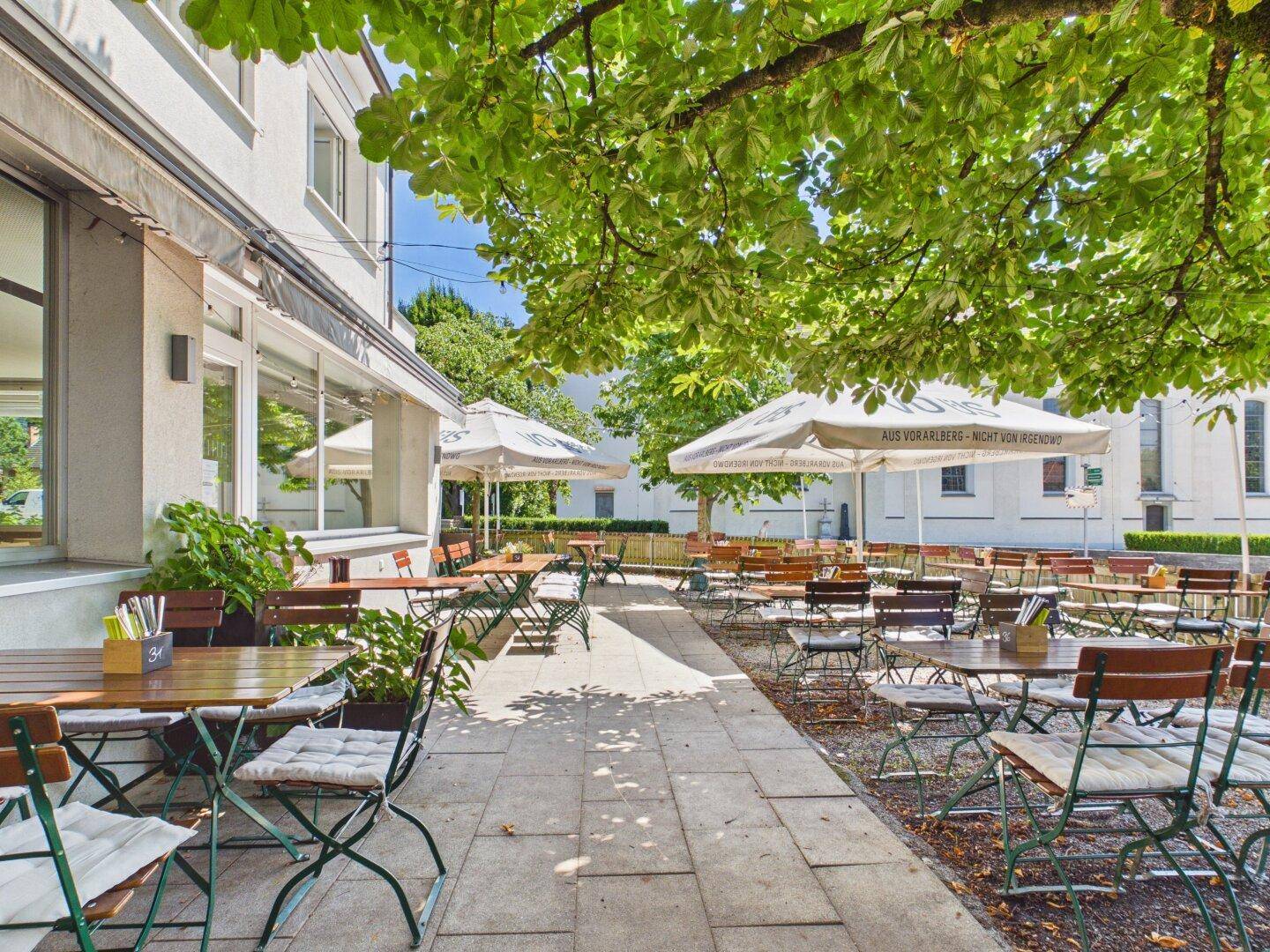 Eine lange, schattige Restaurantterrasse mit vielen Sitzgelegenheiten und Blick auf die umliegende Grünfläche.