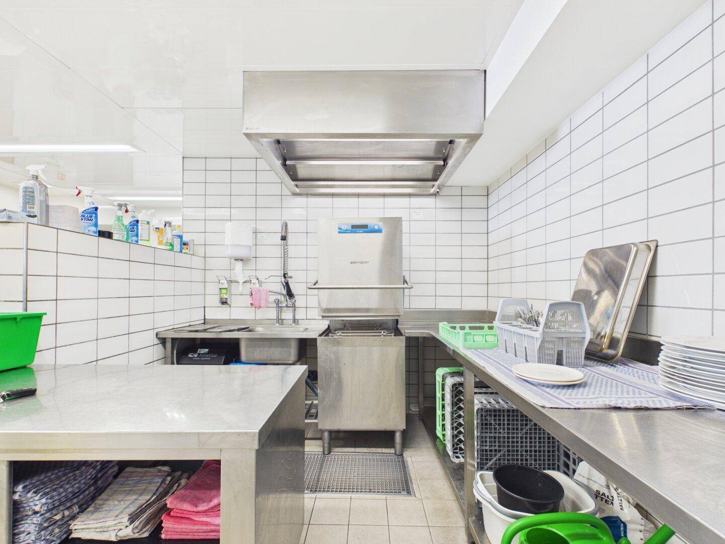 Spülbereich der Großküche mit industrieller Spülmaschine, großem Waschbecken und Ablageflächen.