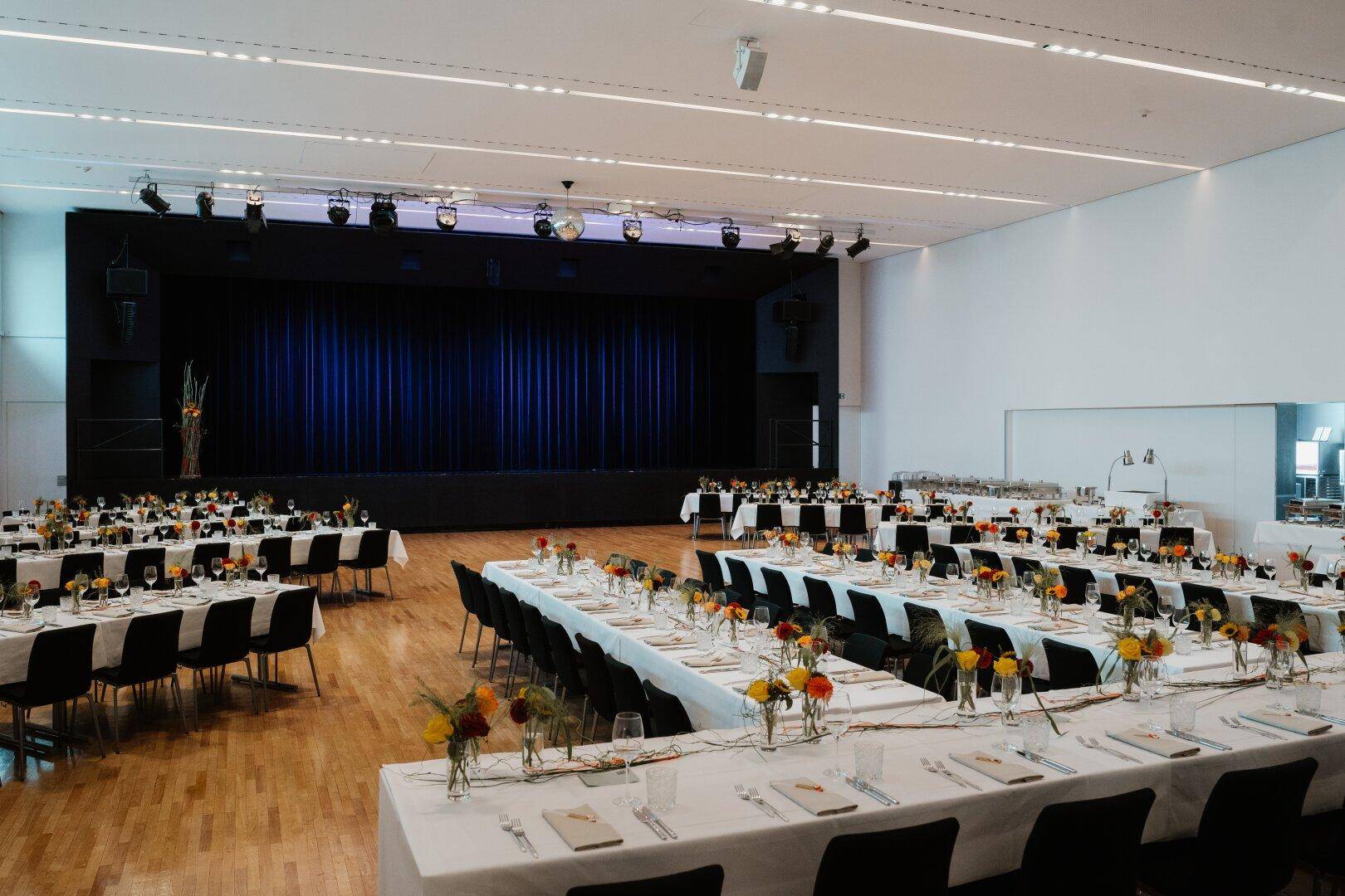 Großer Veranstaltungssaal, festlich gedeckt mit Tischen und Stühlen, bereit für ein Bankett vor einer Bühne.