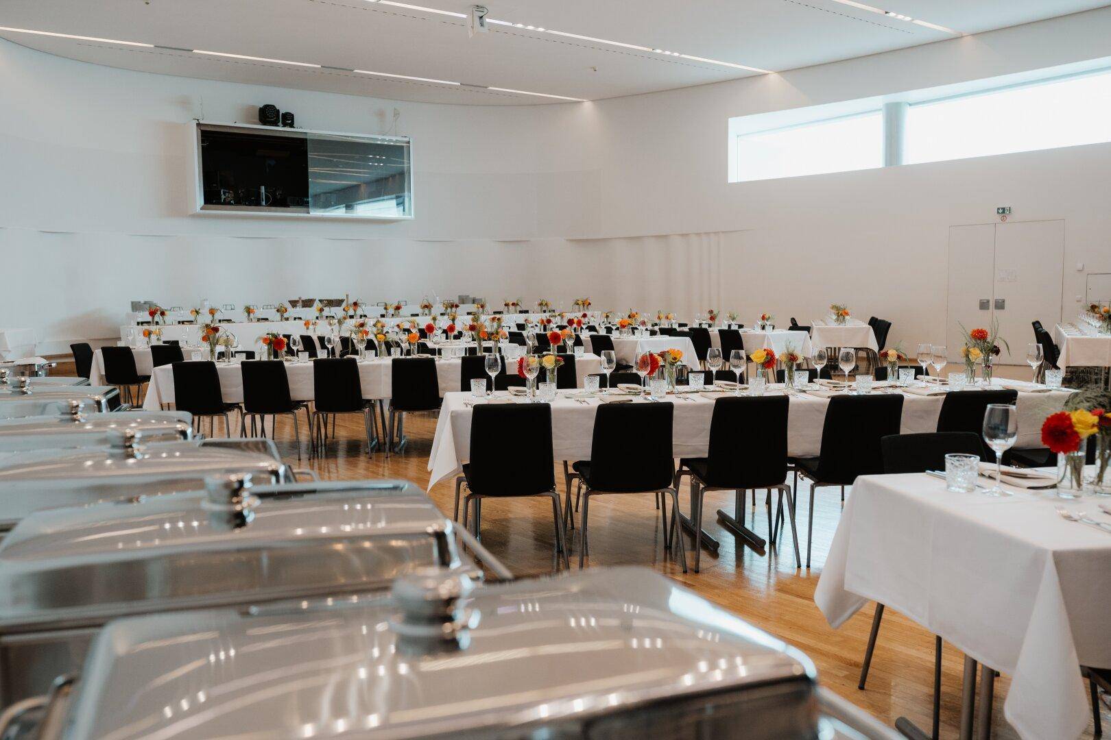 Veranstaltungssaal mit gedeckten Tischen und Buffetwärmern im Vordergrund, bereit für die Bewirtung der Gäste.