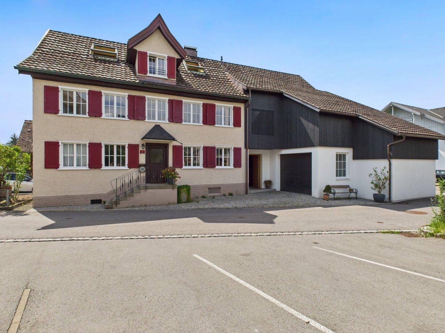 Traditionelles Wohnhaus mit roten Fensterläden und angrenzendem modernen Anbau mit Garage.