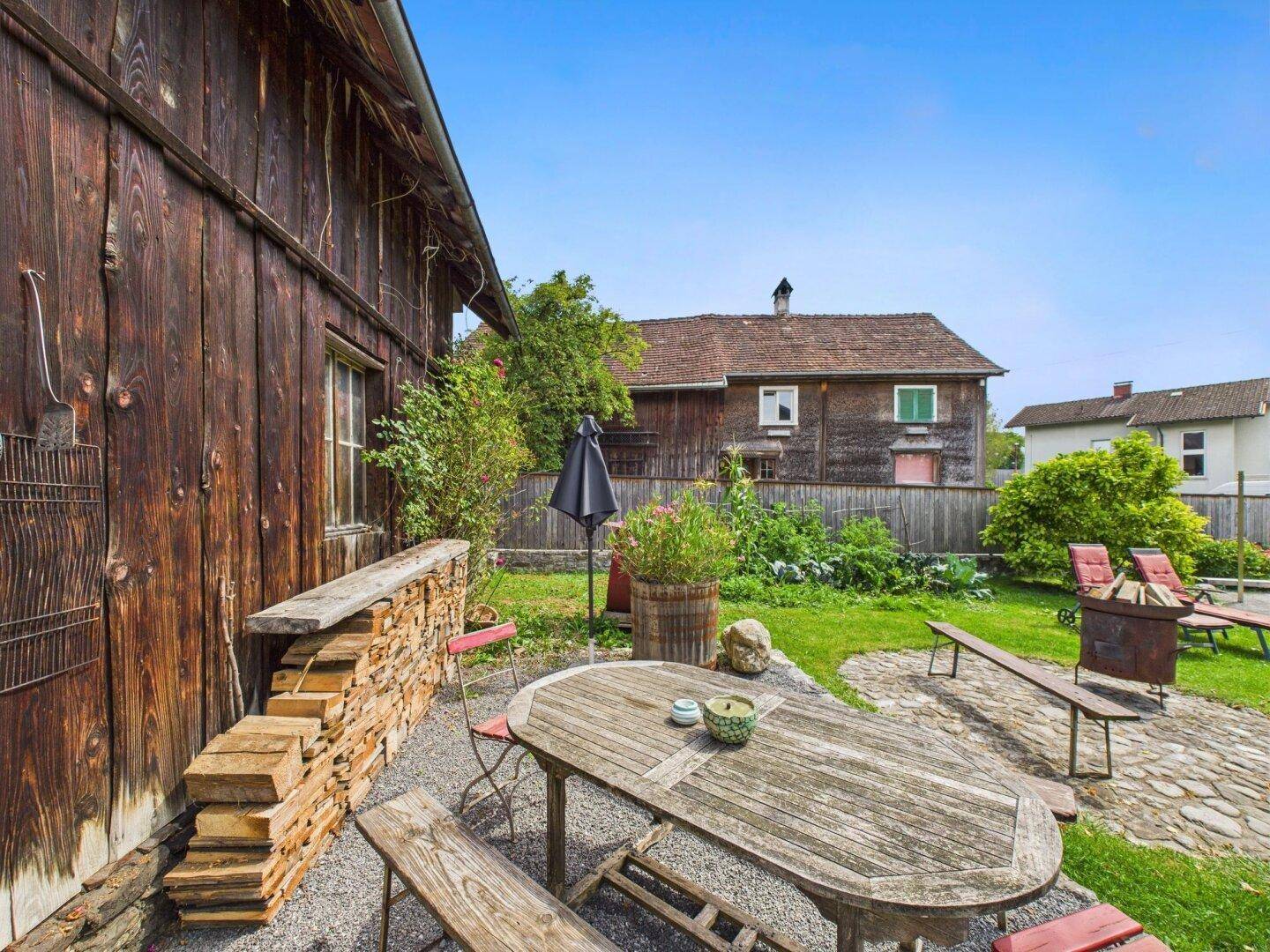 Rustikale Terrasse mit Holztisch, Sitzbänken und Feuerstelle, umgeben von viel Grün.