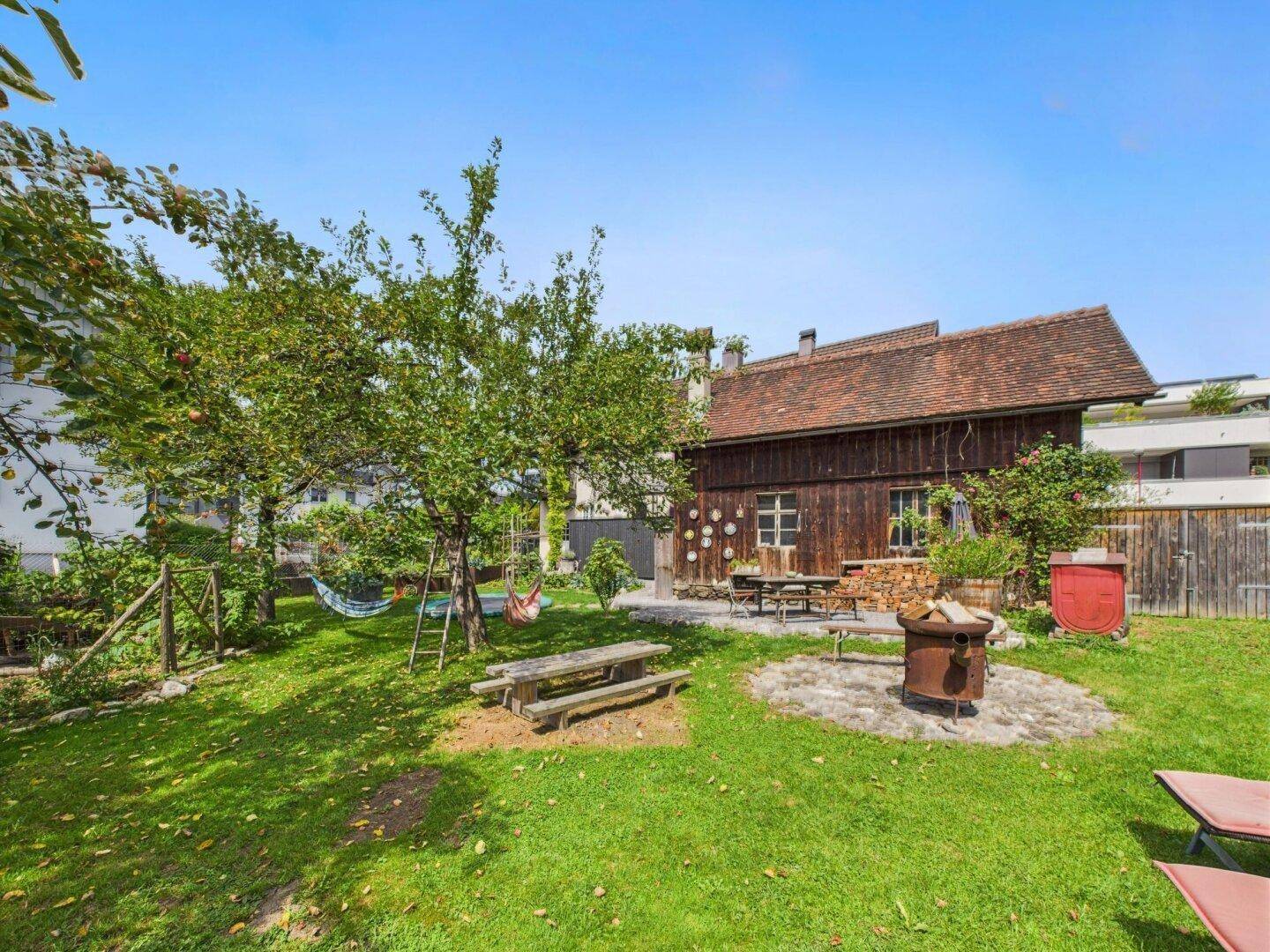 Weitläufiger Garten mit Obstbäumen, Rasenfläche, Feuerstelle und rustikalen Sitzgelegenheiten.