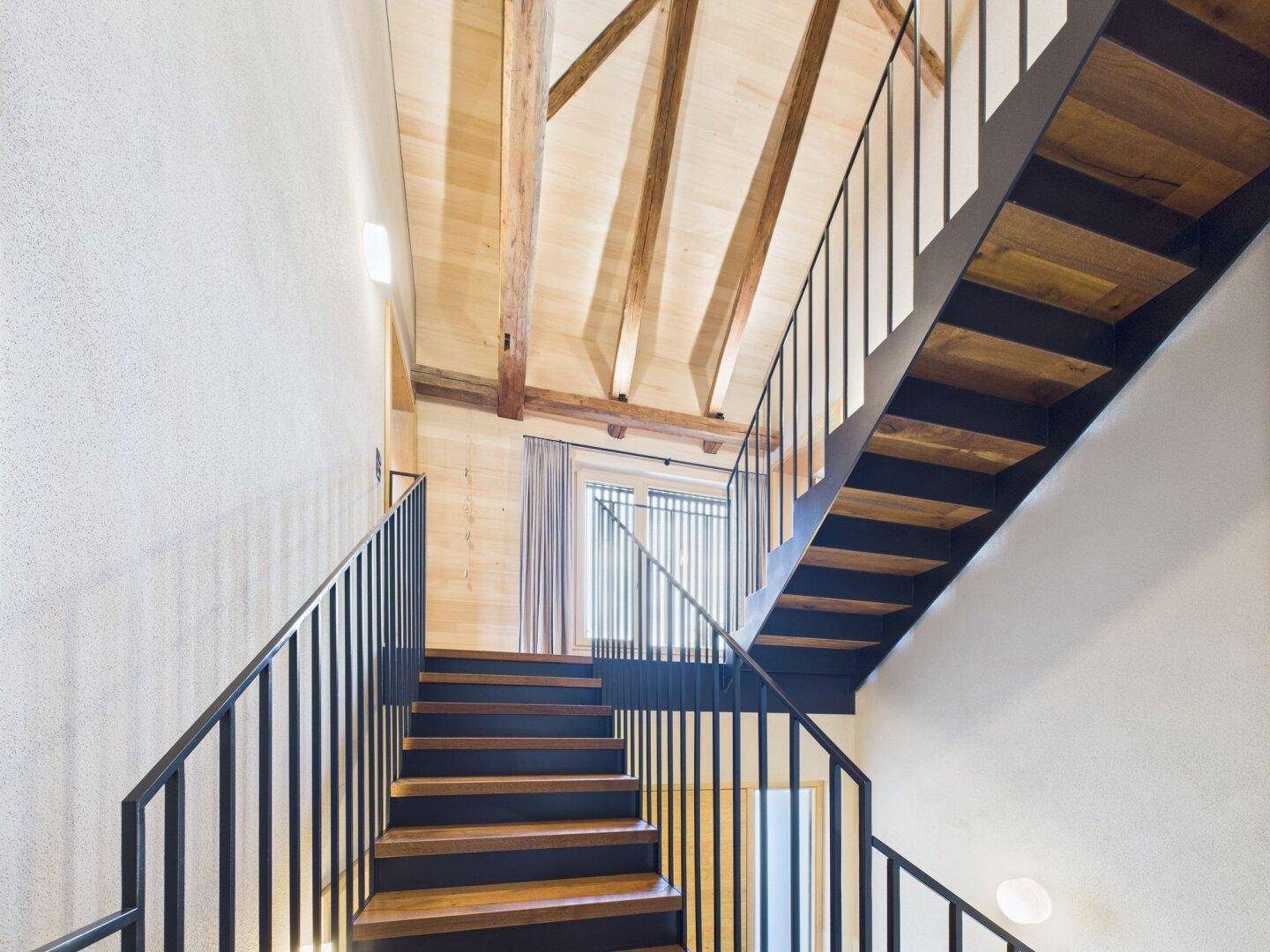 Moderne Holztreppe mit schwarzem Metallgeländer, die zu einem oberen Stockwerk führt.
