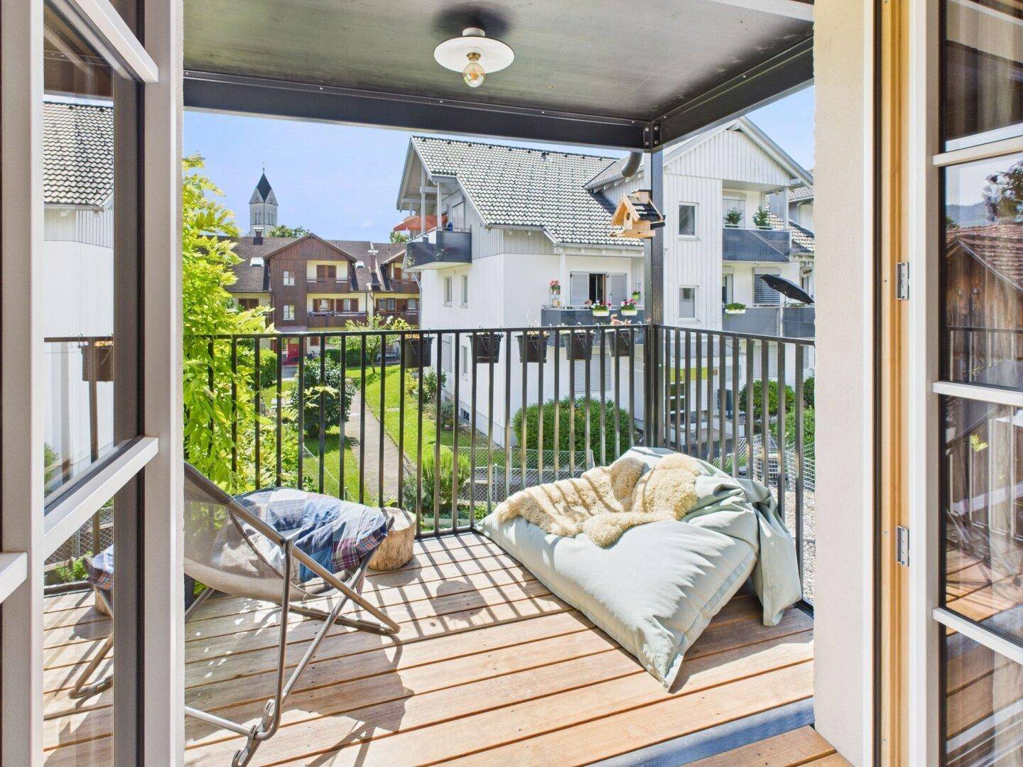 Sonniger Balkon mit Holzbelag, Sitzgelegenheiten und Blick auf die umliegenden Häuser und den Kirchturm.