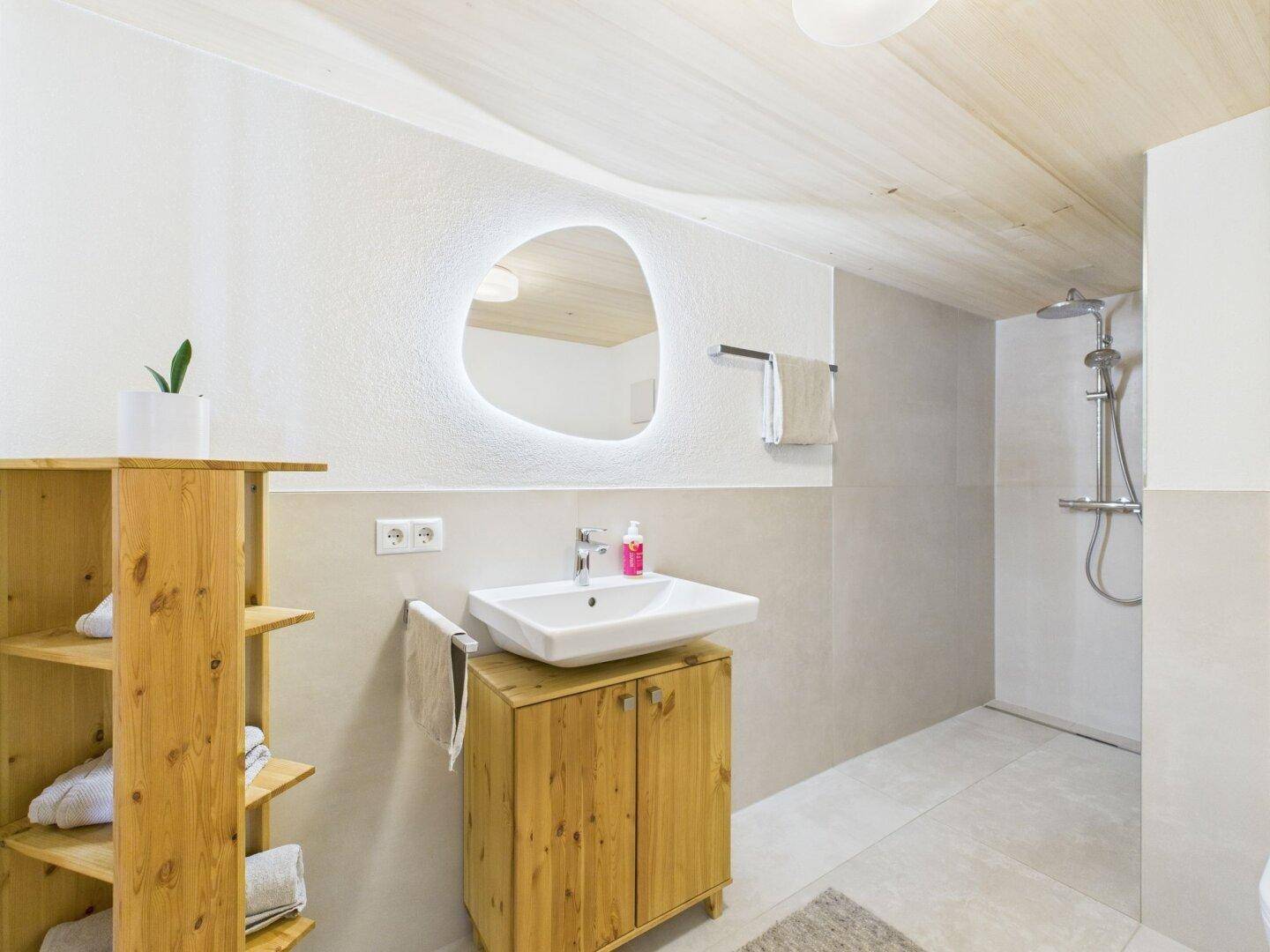 Helles Badezimmer mit beleuchtetem Spiegel, Waschbecken und Duschbereich.