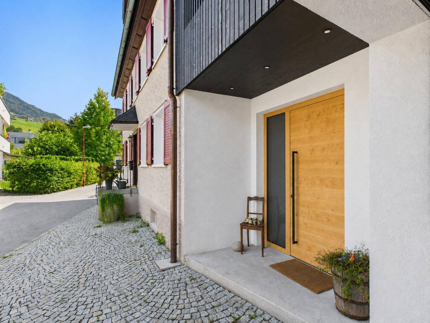 Modernes Haus mit Holztür, gepflastertem Eingangsbereich und Blick auf grüne Hügel.