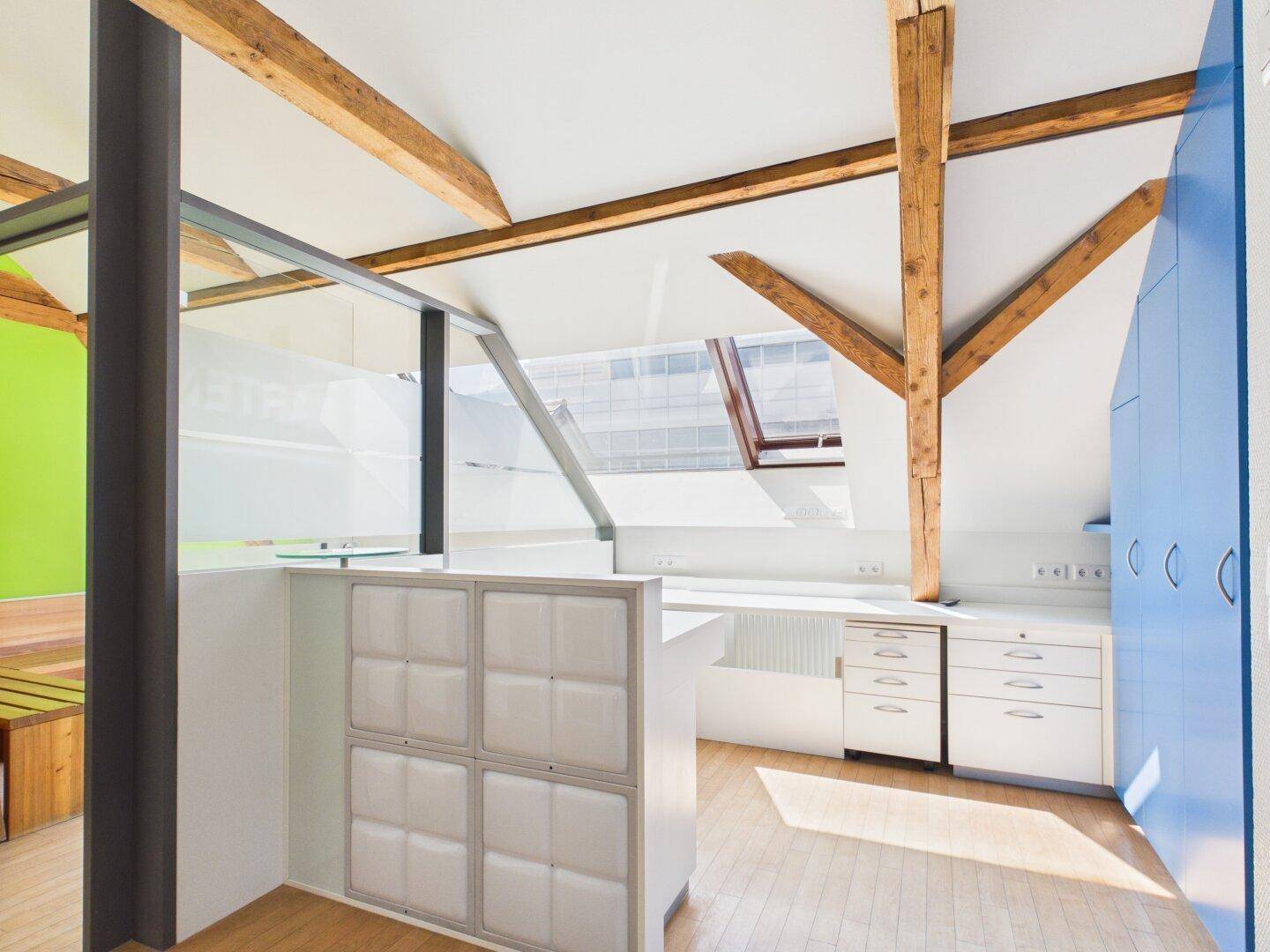 Modernes Büro mit Holzbalken, Dachfenstern und blauen Einbauschränken.