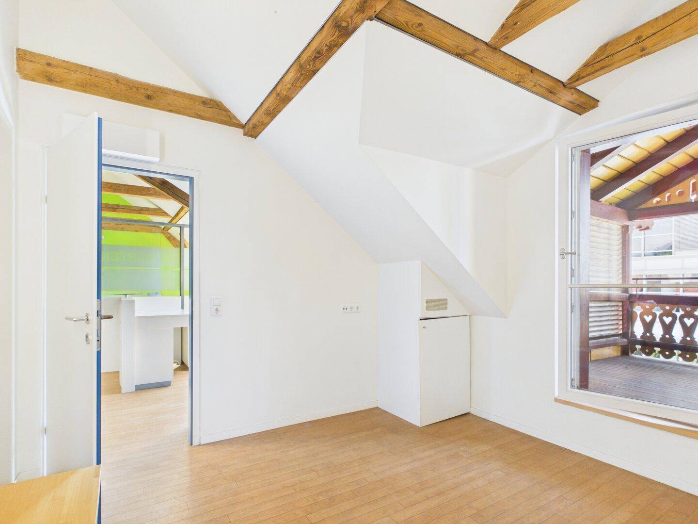Heller Büroraum mit Holzboden, Fenster und Zugang zu einem Balkon.