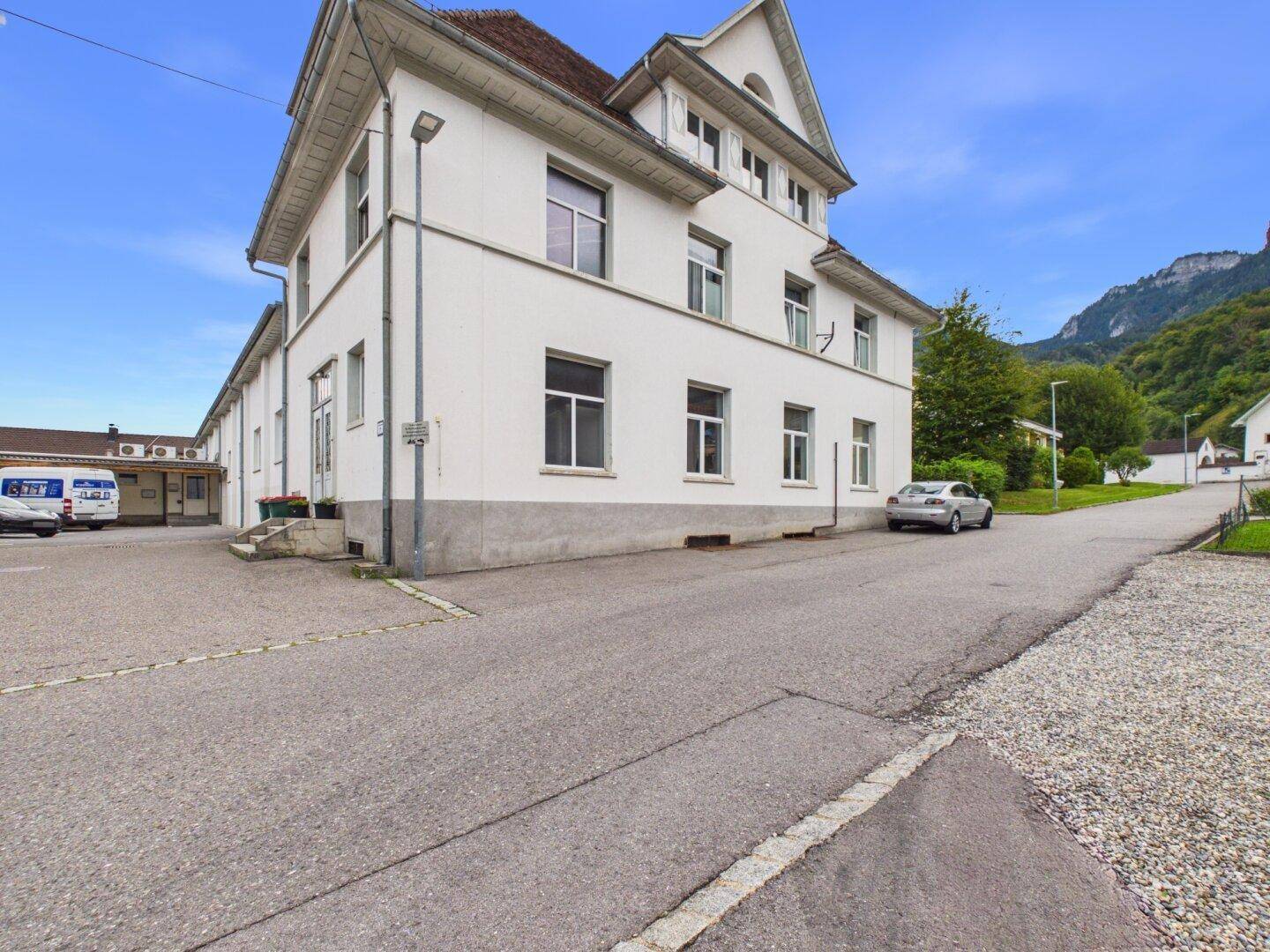 Weißes Gebäude mit traditioneller Architektur und Bergblick, umgeben von einer asphaltierten Zufahrt.