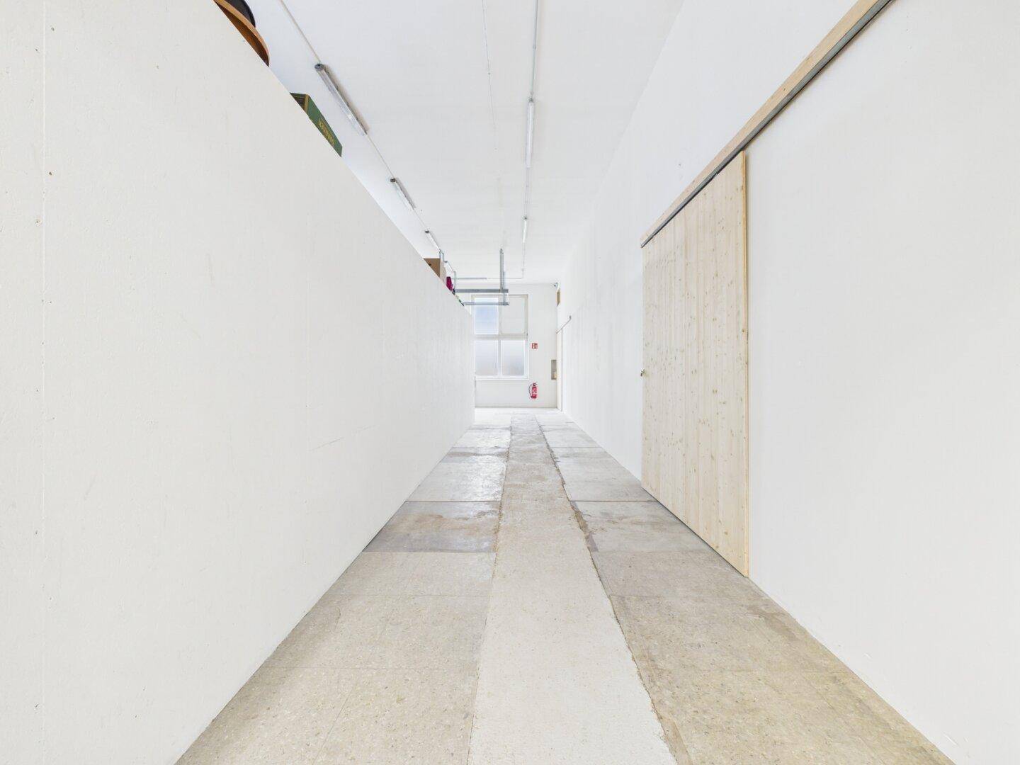 Langer, schlichter Korridor mit weißen Wänden und Betonboden, ausgestattet mit einer Holzschiebetür.