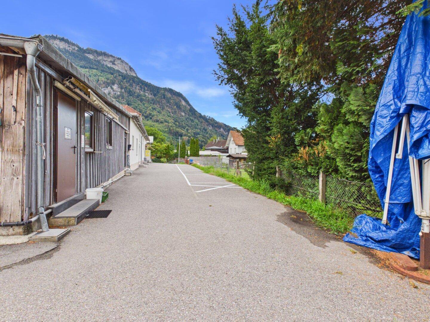 Asphaltierte Zufahrt mit einem Holzgebäude auf der linken Seite und einer grünen Berglandschaft im Hintergrund.