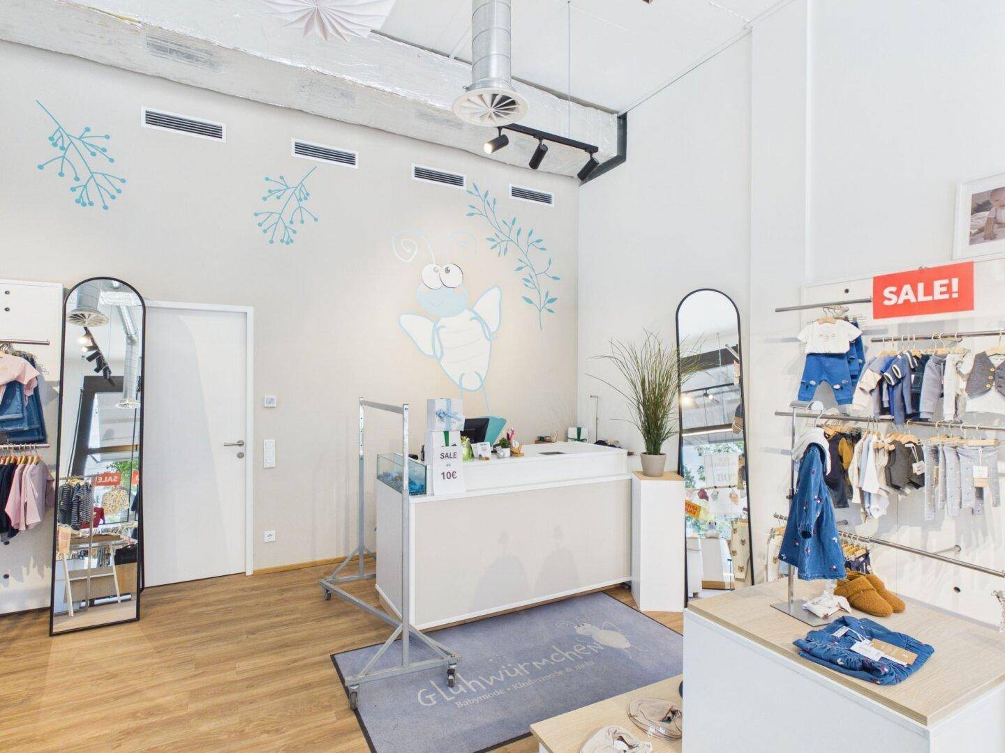 Kassenbereich eines Kinderbekleidungsgeschäfts mit heller Theke, Wanddekoration und großen Spiegeln.