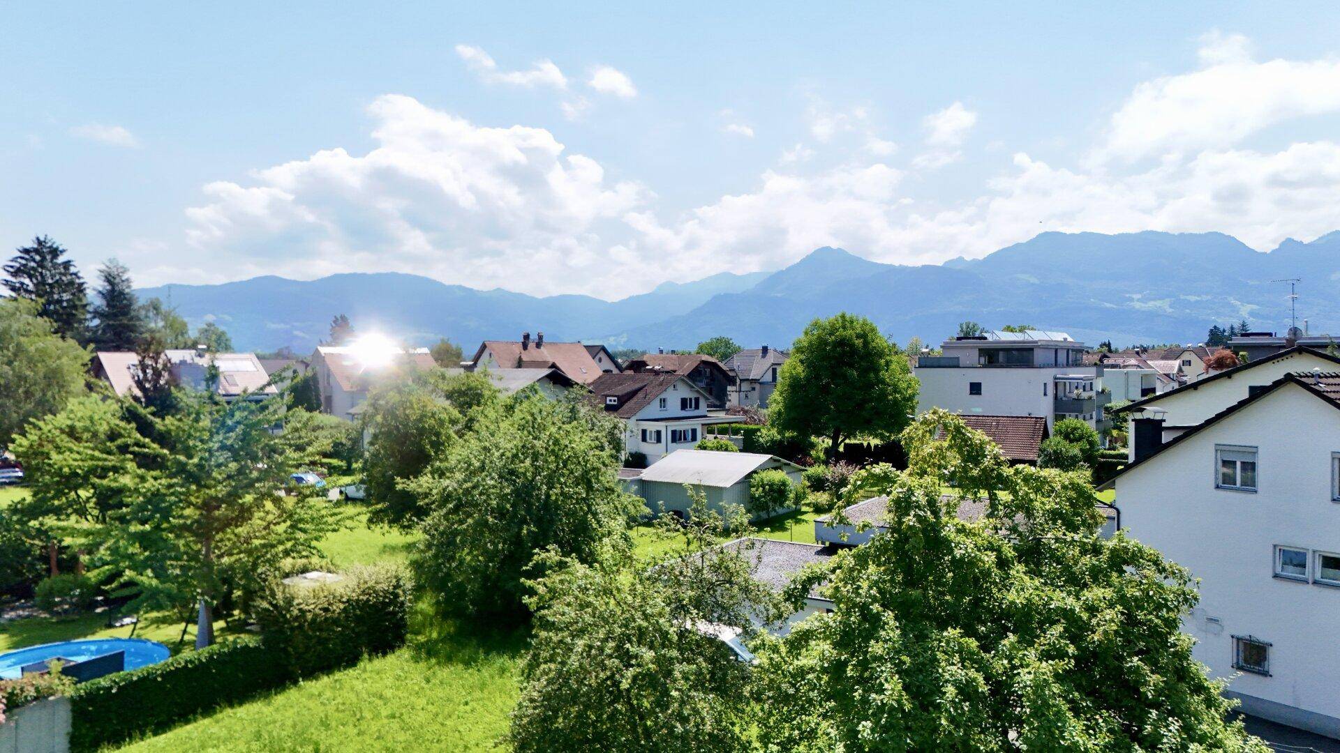 Panoramablick über eine grüne Wohnsiedlung mit Häusern und majestätischen Bergen im Hintergrund unter blauem Himmel.