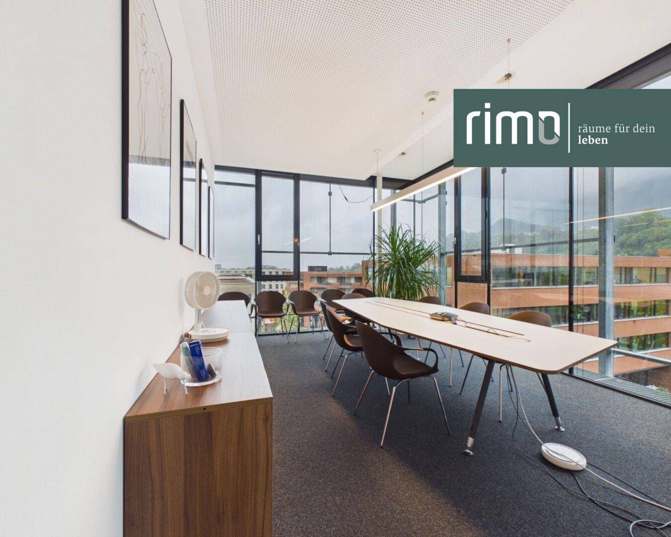 Heller Konferenzraum mit Teppichboden und einer langen Tafel, umgeben von großen Fenstern.