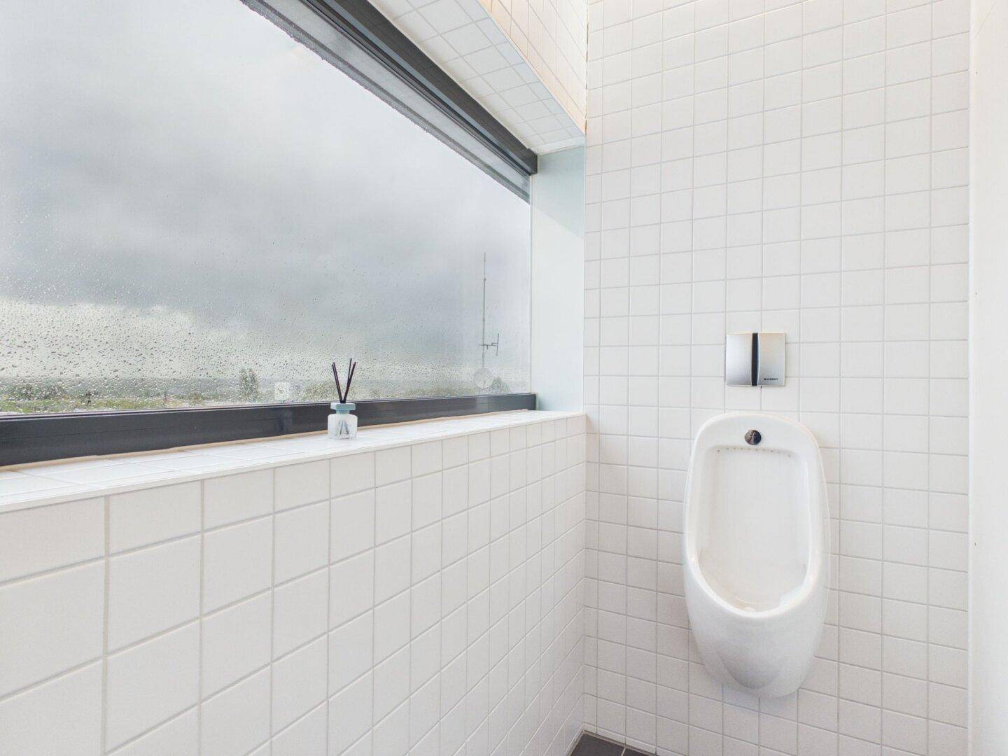 Toilettenraum mit Urinal, weißen Fliesen und einem Fenster für Tageslicht.