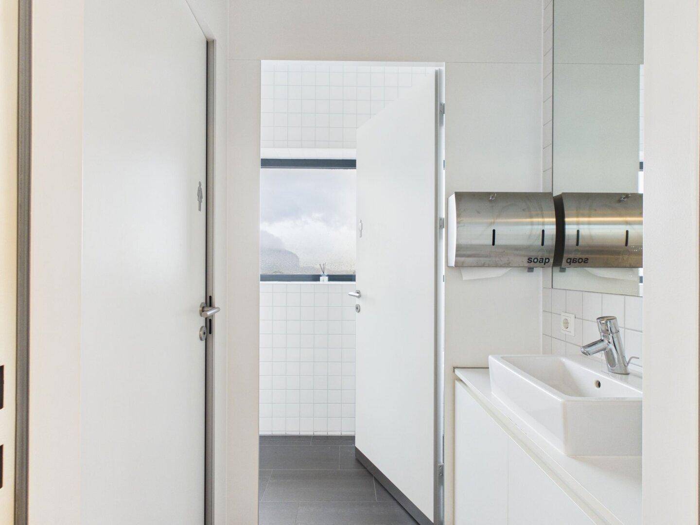 Heller Toilettenbereich mit Waschbecken, Seifenspender und Zugang zu weiteren Kabinen.