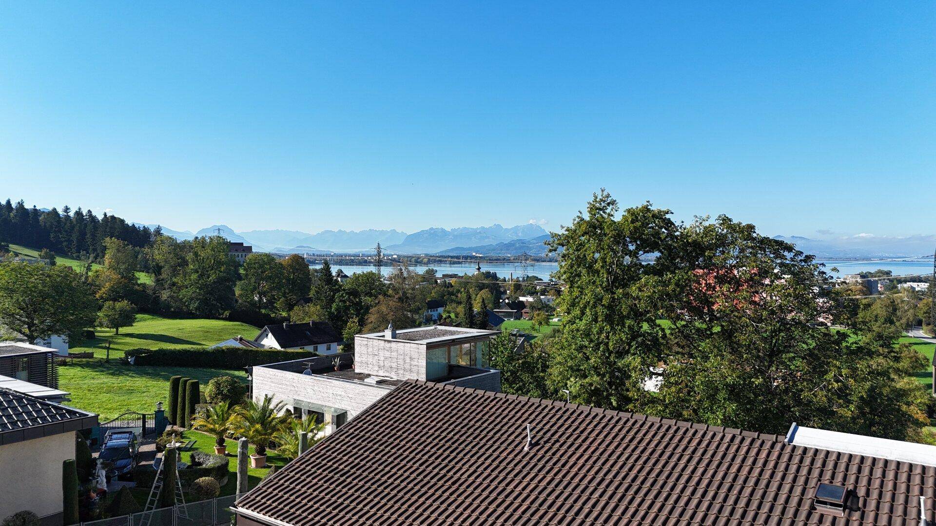Weitläufiger Blick über eine Wohngegend mit Dächern, See und Alpenpanorama unter blauem Himmel.