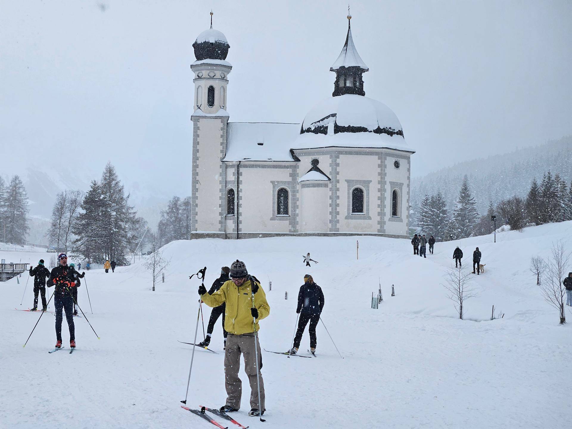 Langläufer gleiten durch eine verschneite Landschaft vor einer großen Kirche mit Zwiebeltürmen.