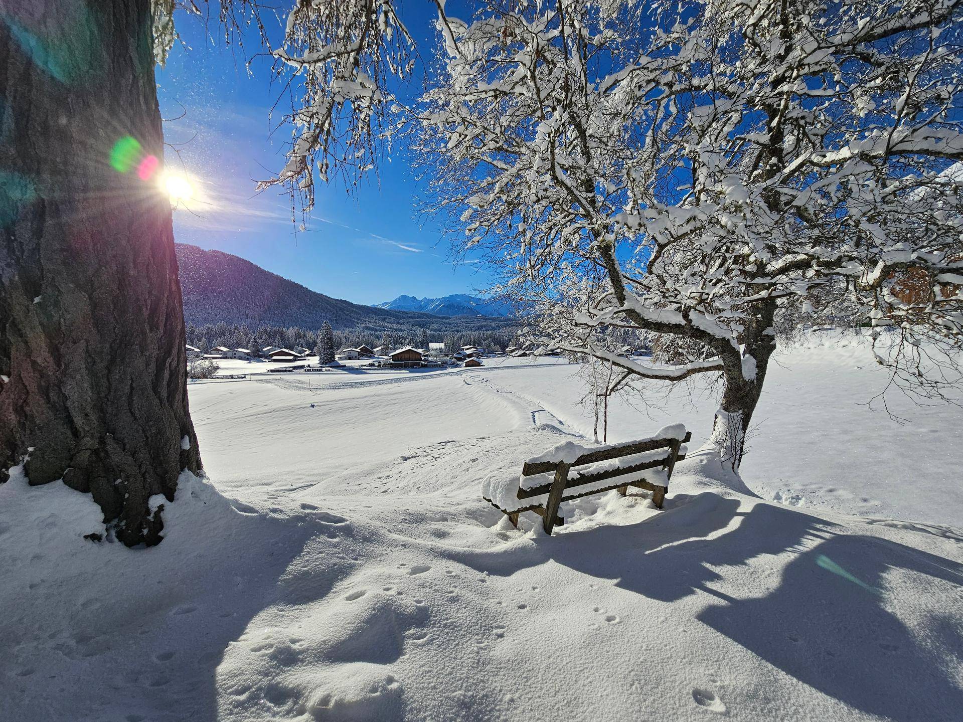 Eine verschneite Holzbank unter einem Baum bietet einen malerischen Blick auf ein Winterdorf und ferne Berge.
