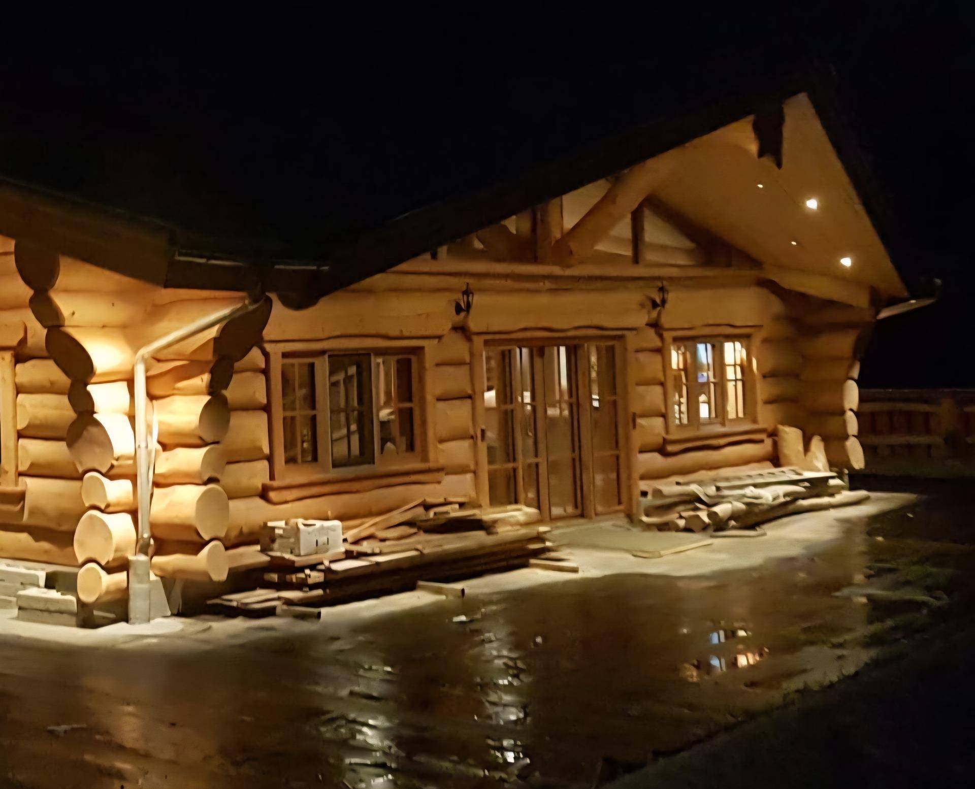 Blockhausfassade bei Nacht mit stimmungsvoller Beleuchtung und Baumaterialien im Vordergrund.
