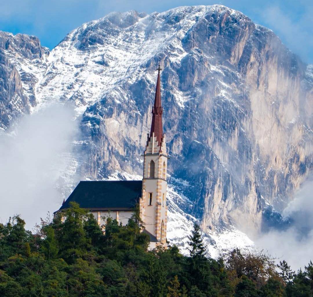 Historische Kirche auf einem Hügel vor einer beeindruckenden, schneebedeckten Bergkulisse.