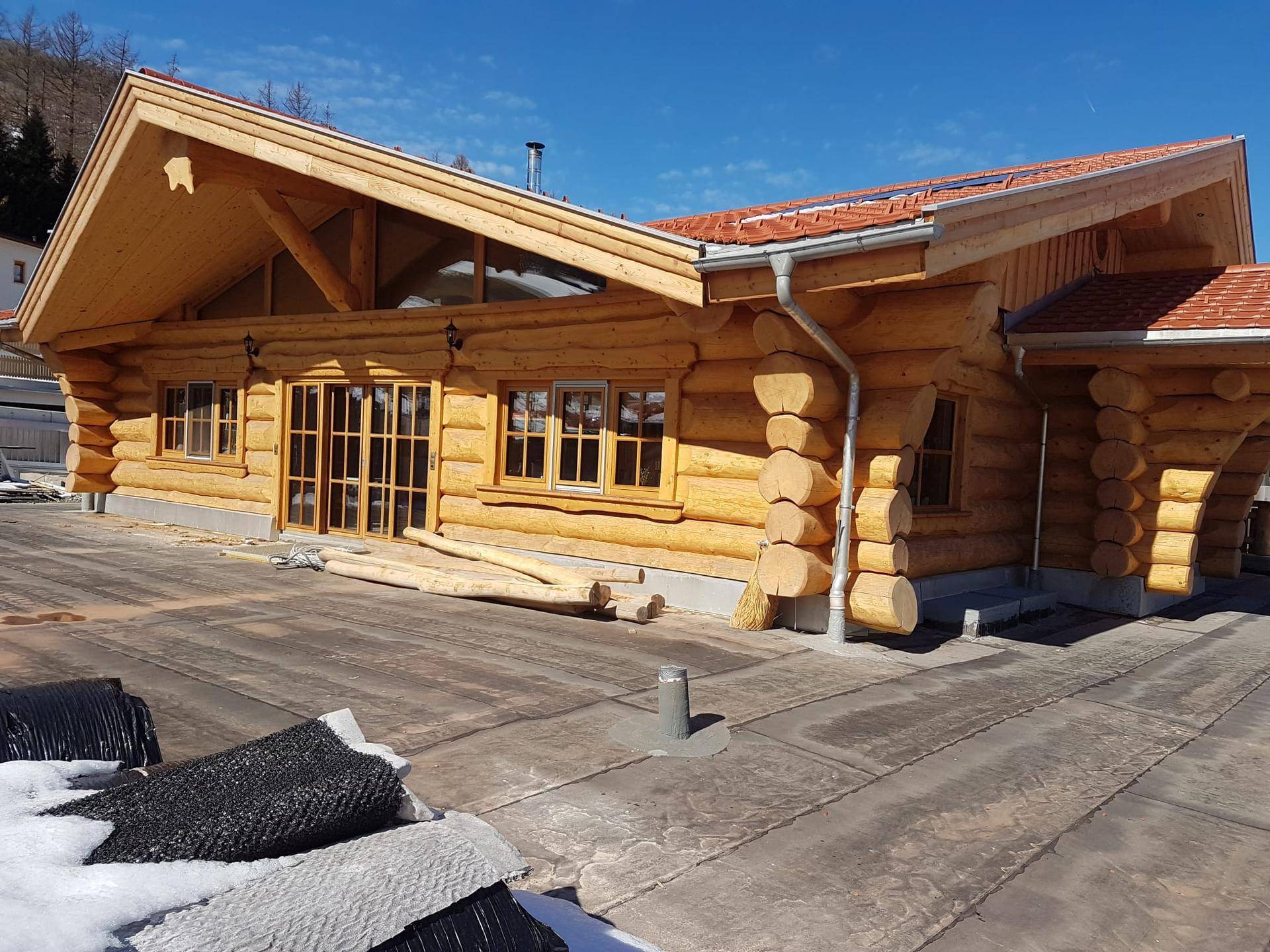 Blockhaus im Bau mit Holzbalken, großen Fenstern und Baustelle im Vordergrund an einem sonnigen Tag.