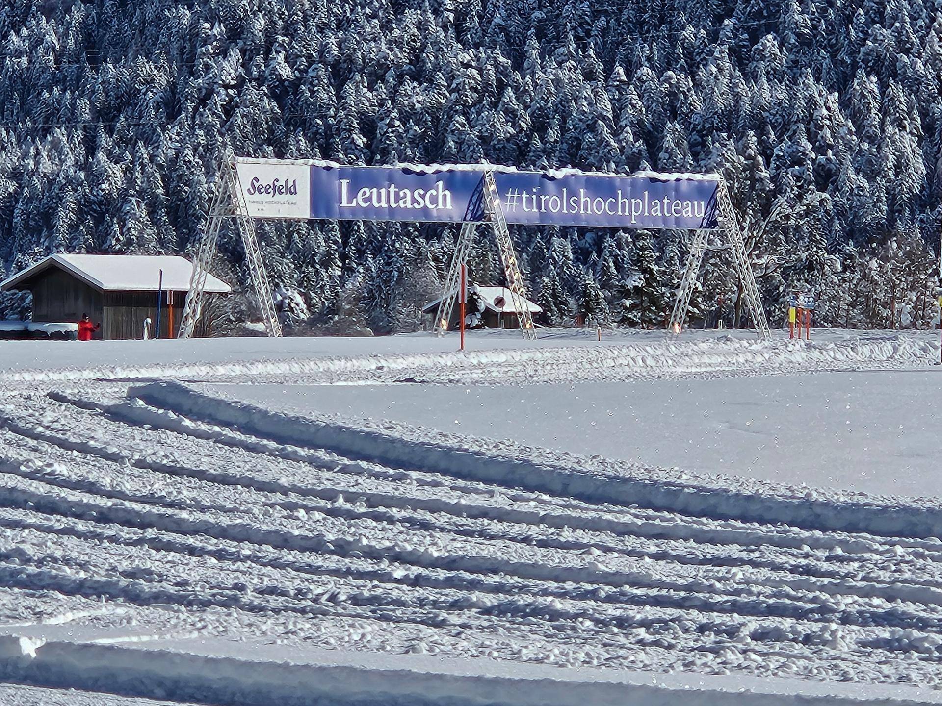 Eine präparierte Skipiste führt durch einen verschneiten Wald mit einem Werbebanner im Hintergrund.