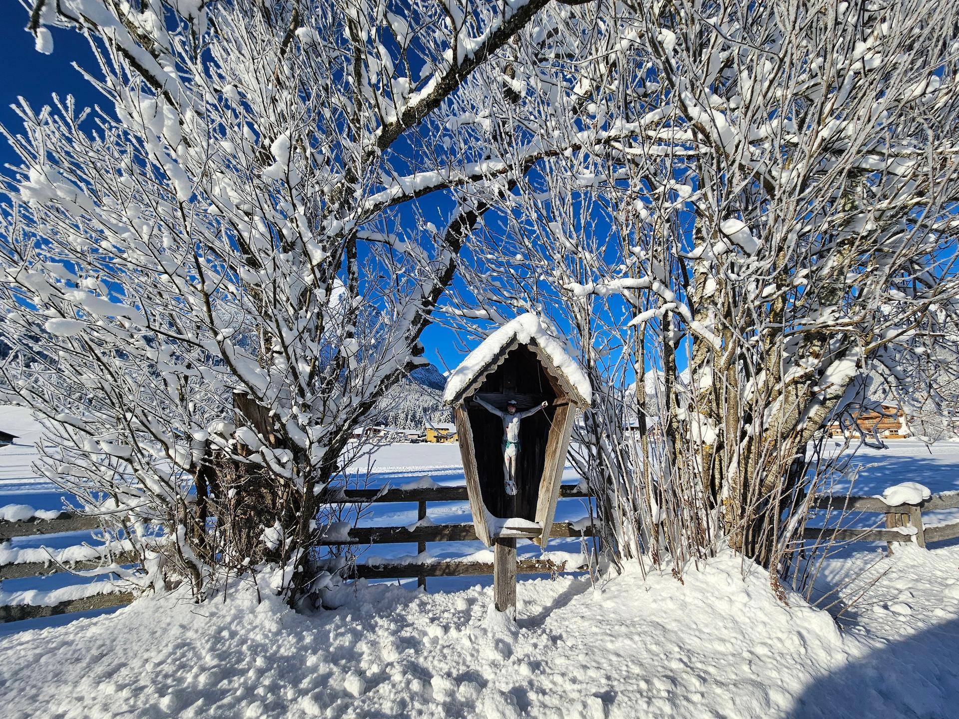 Ein kleines Holzkreuz mit Kruzifix steht umgeben von schneebedeckten Bäumen in einer winterlichen Landschaft.