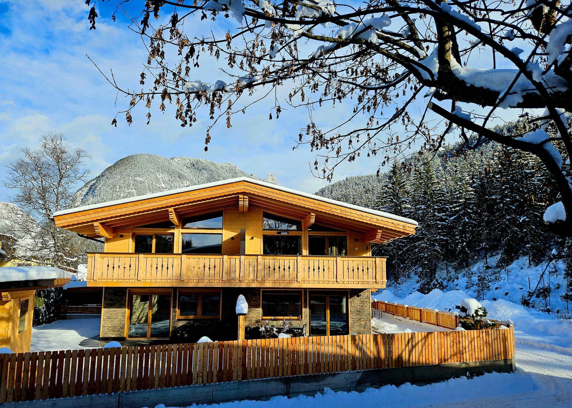 Modernes Holzhaus im Winter mit Balkon und verschneiter Umgebung vor bewaldeten Bergen.