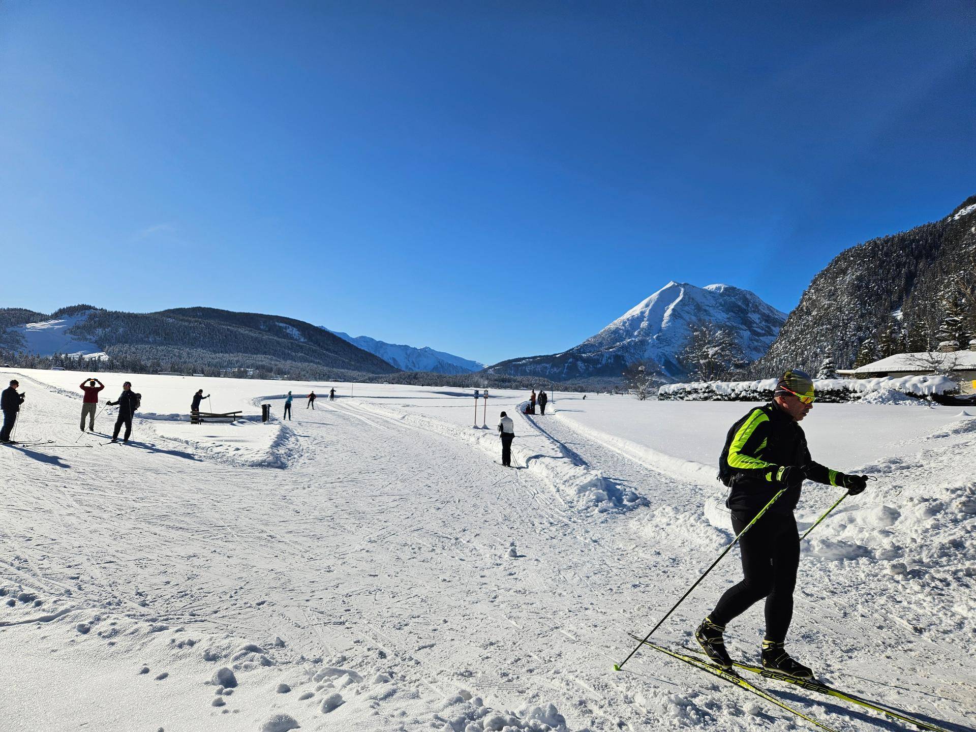 Langläufer auf einer Loipe in einer weiten, sonnigen Winterlandschaft mit Bergen im Hintergrund.