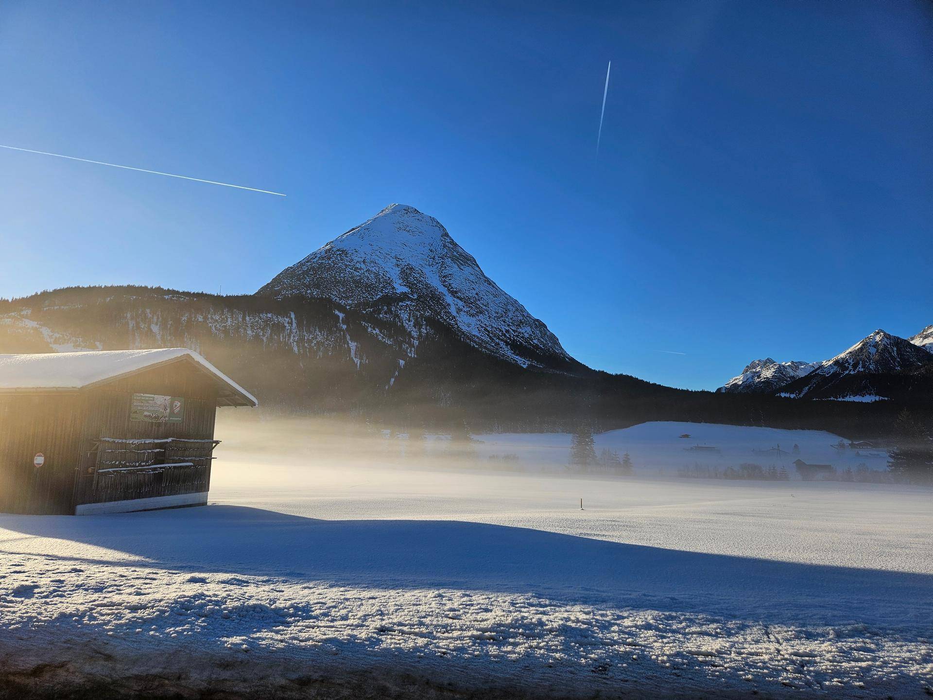 Verschneite Winterlandschaft mit einer Holzhütte und einem majestätischen Berg im Nebel bei Sonnenaufgang.