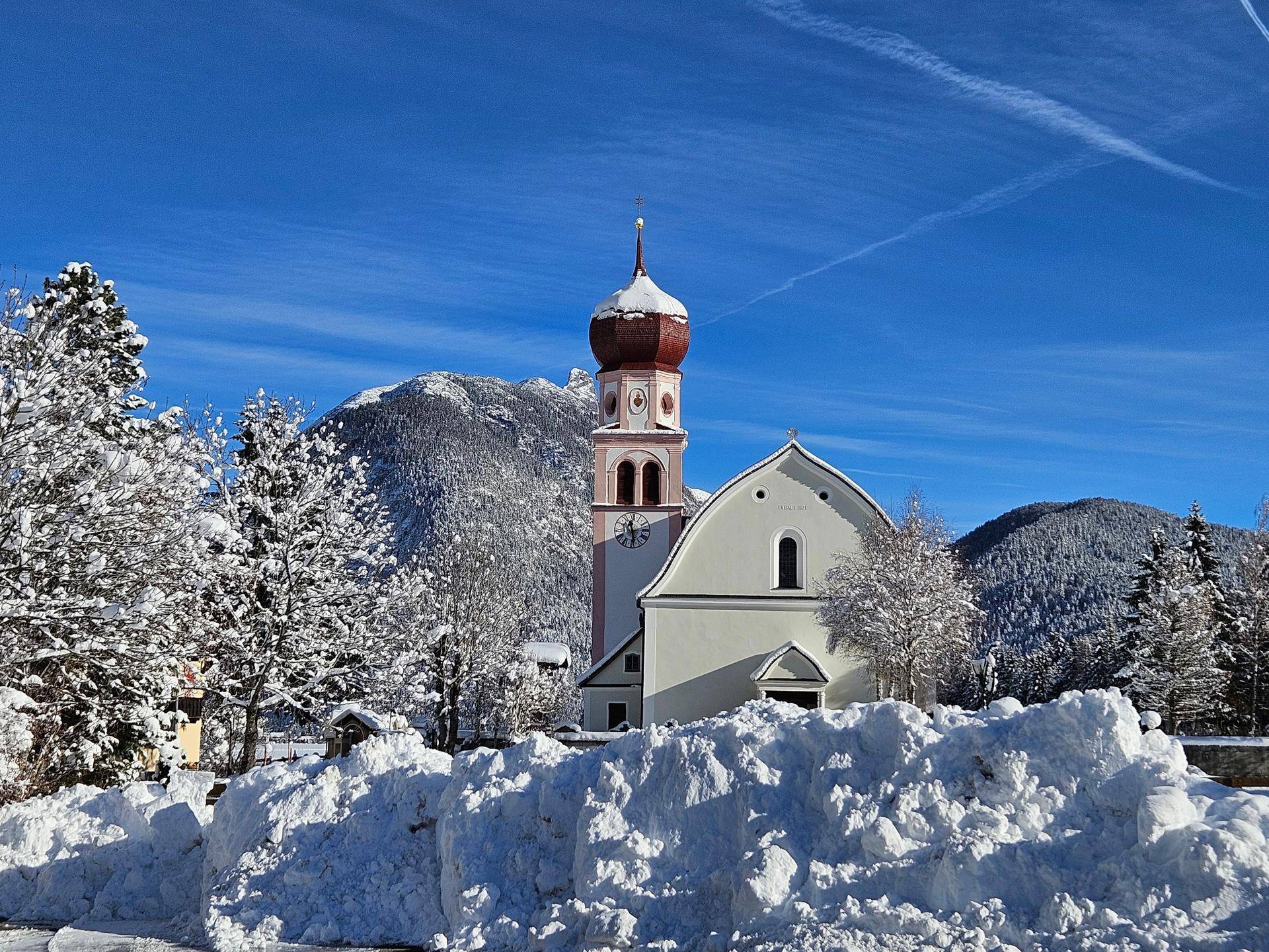 Malerische Kirche in einer tief verschneiten Winterlandschaft mit Bergen unter blauem Himmel.
