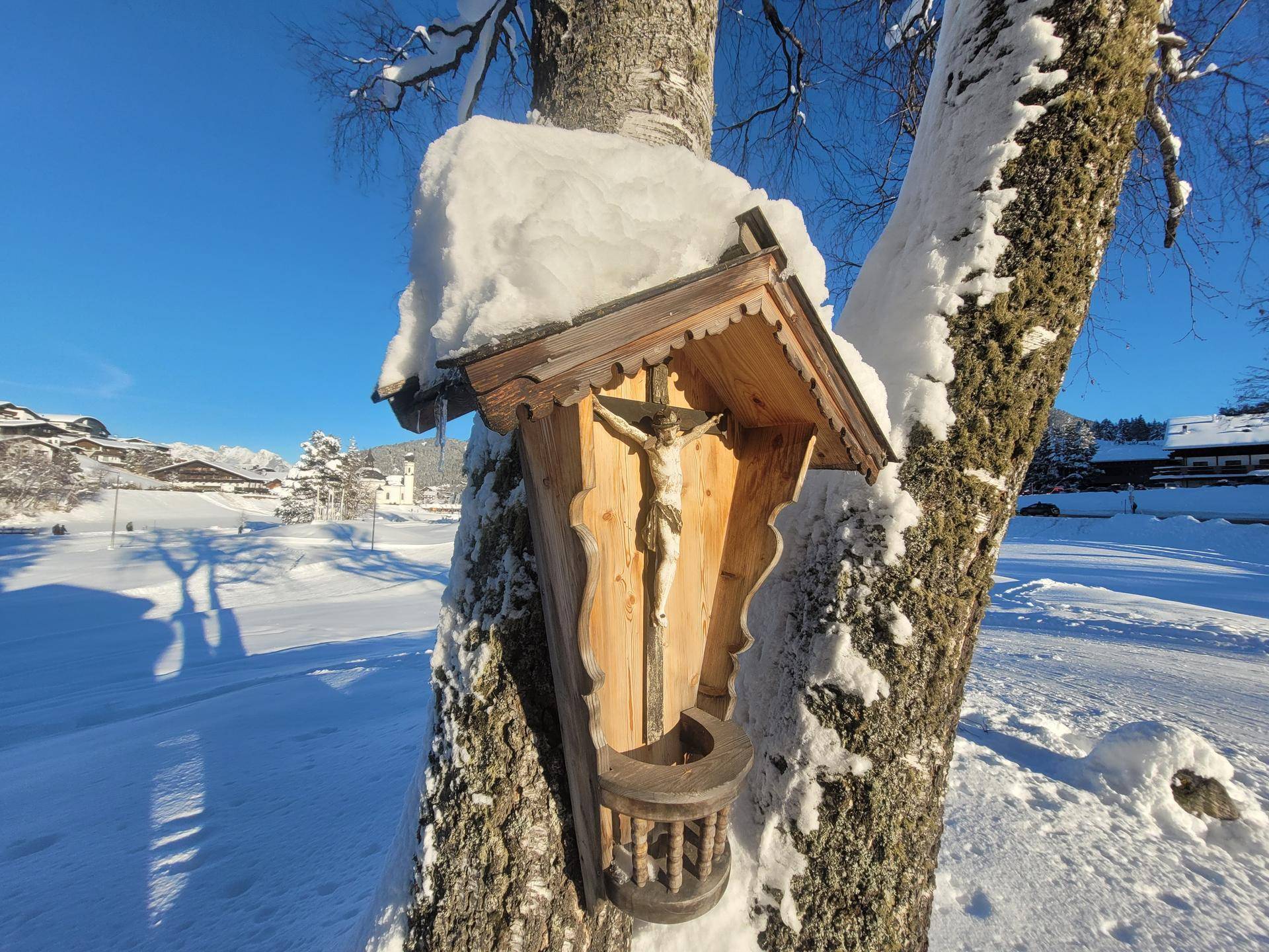 Traditioneller hölzerner Bildstock mit Kruzifix an einem Baum in verschneiter Winterlandschaft.