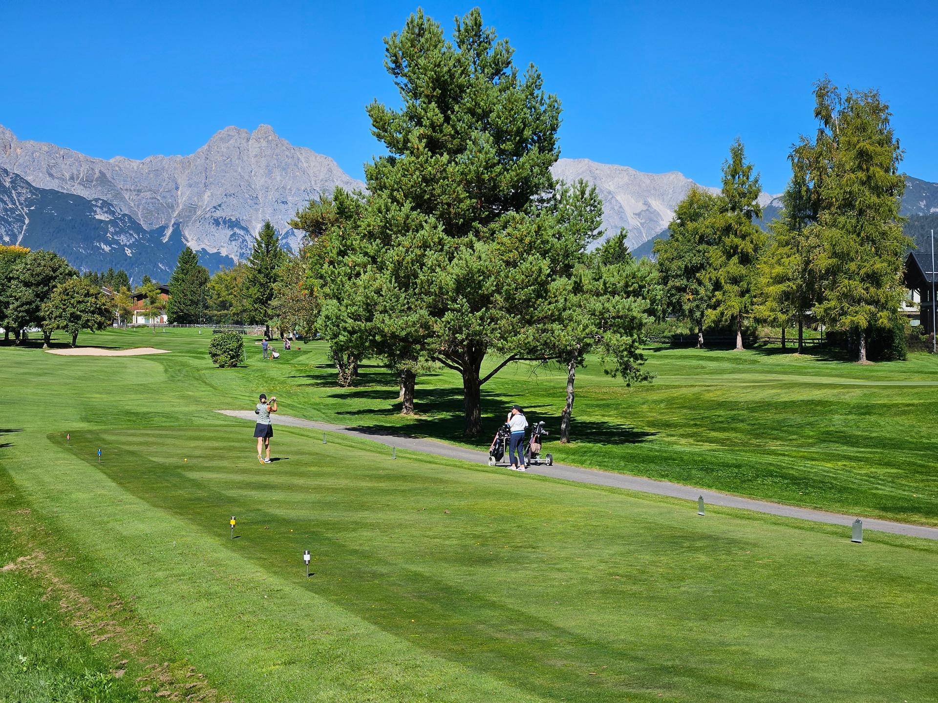 Golfer auf einem grünen Golfplatz mit hohen Bäumen und einer beeindruckenden Bergkulisse im Hintergrund.