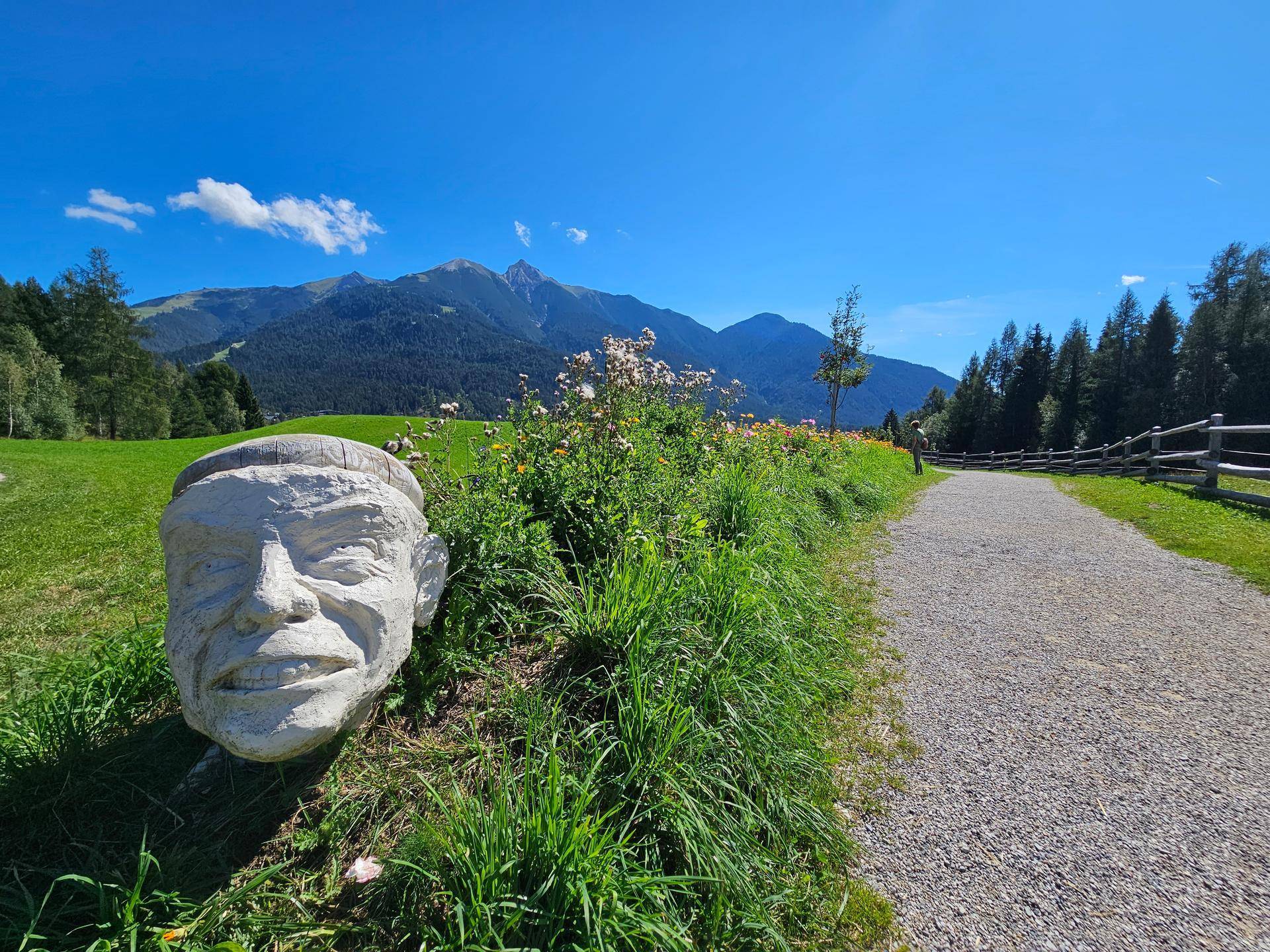 Ein Schotterweg führt durch eine grüne Landschaft mit einer Steinskulptur und Bergen im Hintergrund.